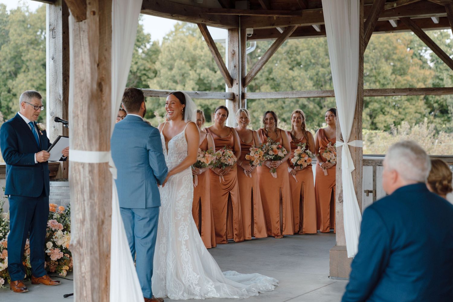 Elegant wedding ceremony taking place under a wooden pavilion with bridesmaids in terracotta dresses standing in a row.