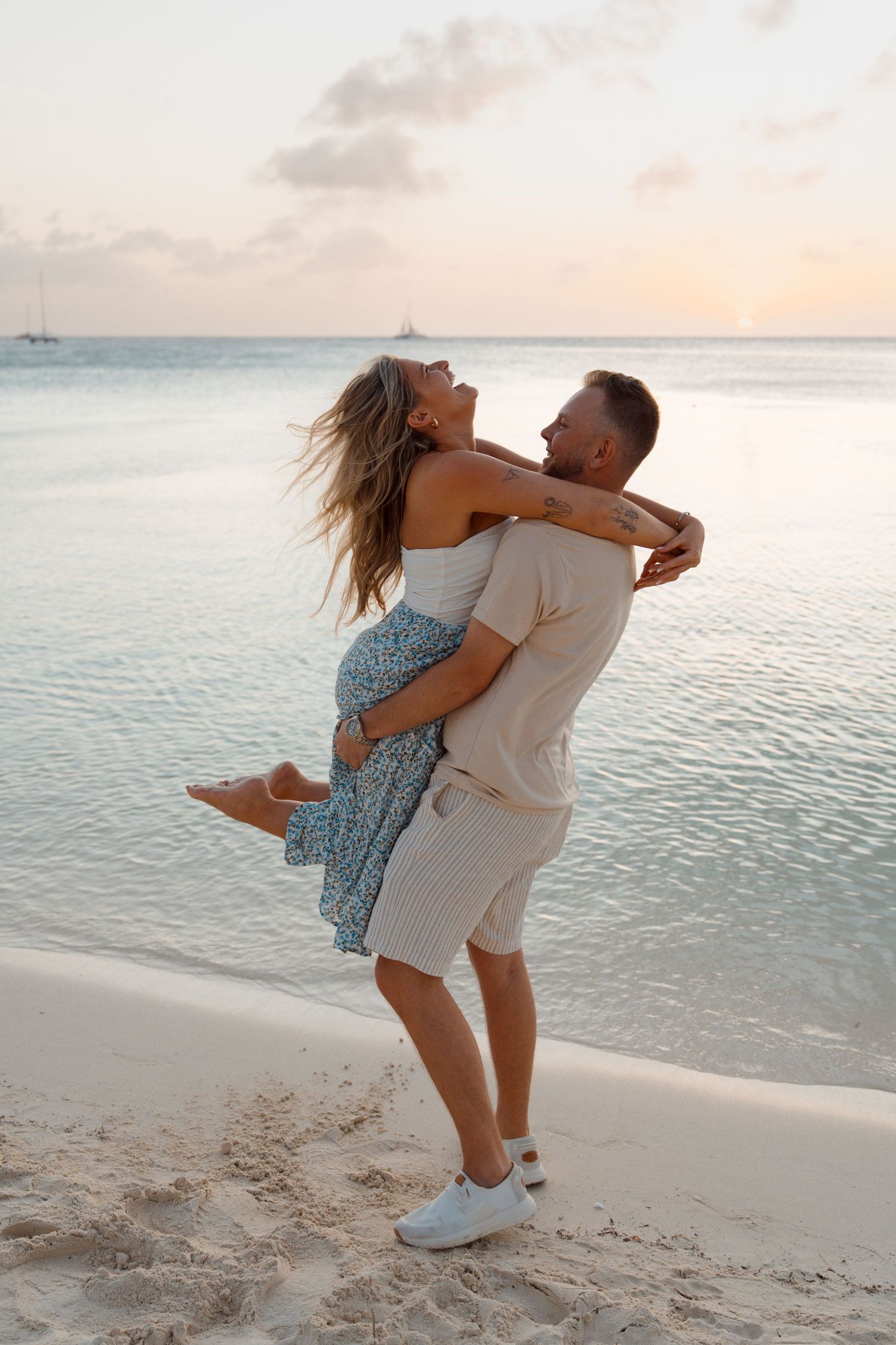 Romantic couple embracing on tropical beach at sunset with turquoise ocean water and pink sky in background.