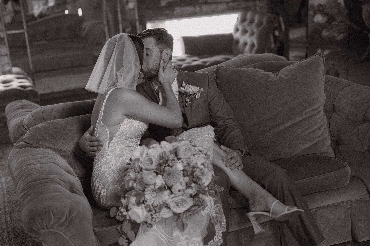 Black and white photo of a bride and groom sharing a kiss on a velvet sofa, bride holding a floral bouquet.