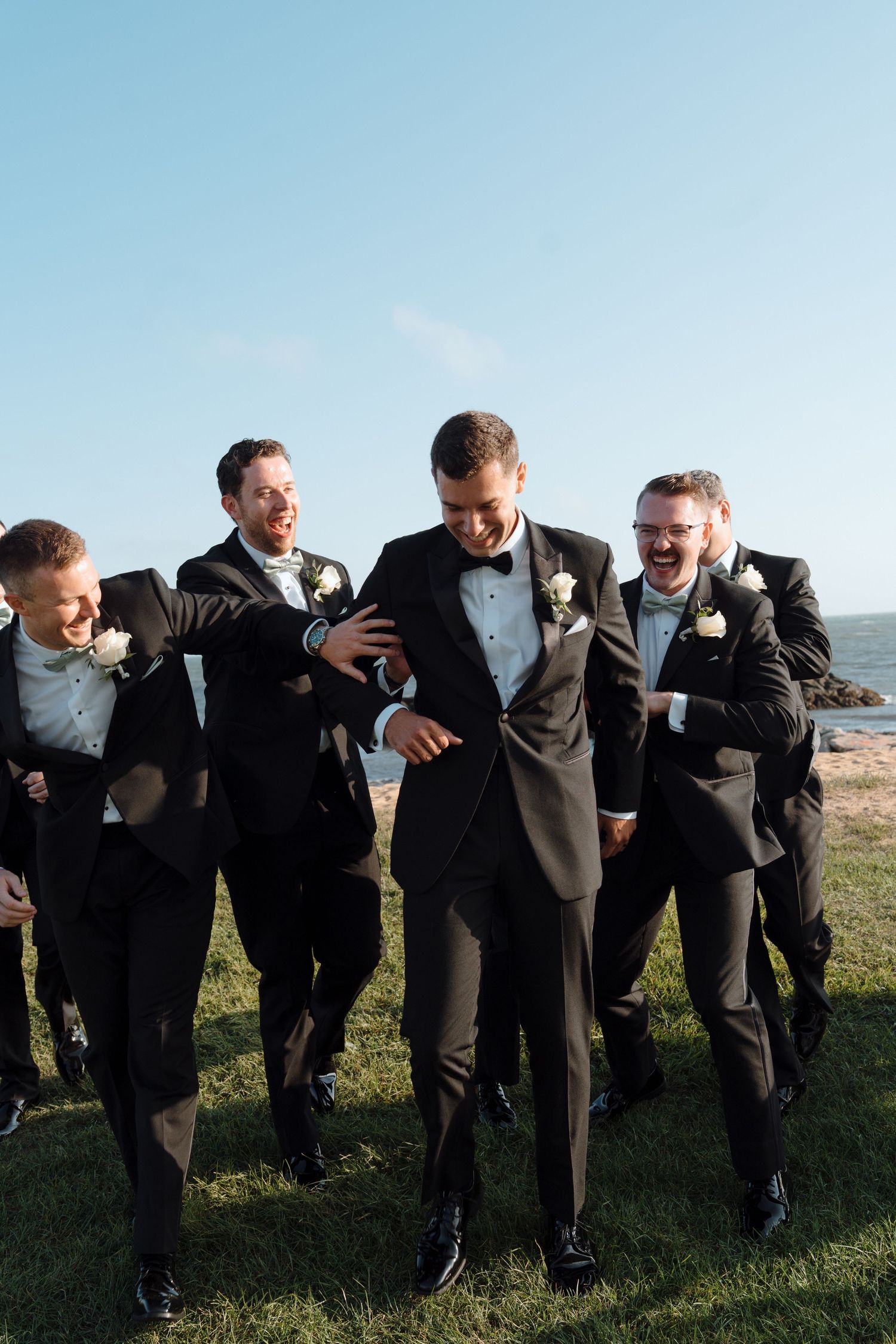 Groom and groomsmen in black suits walking together outdoors on grass with ocean view in background.