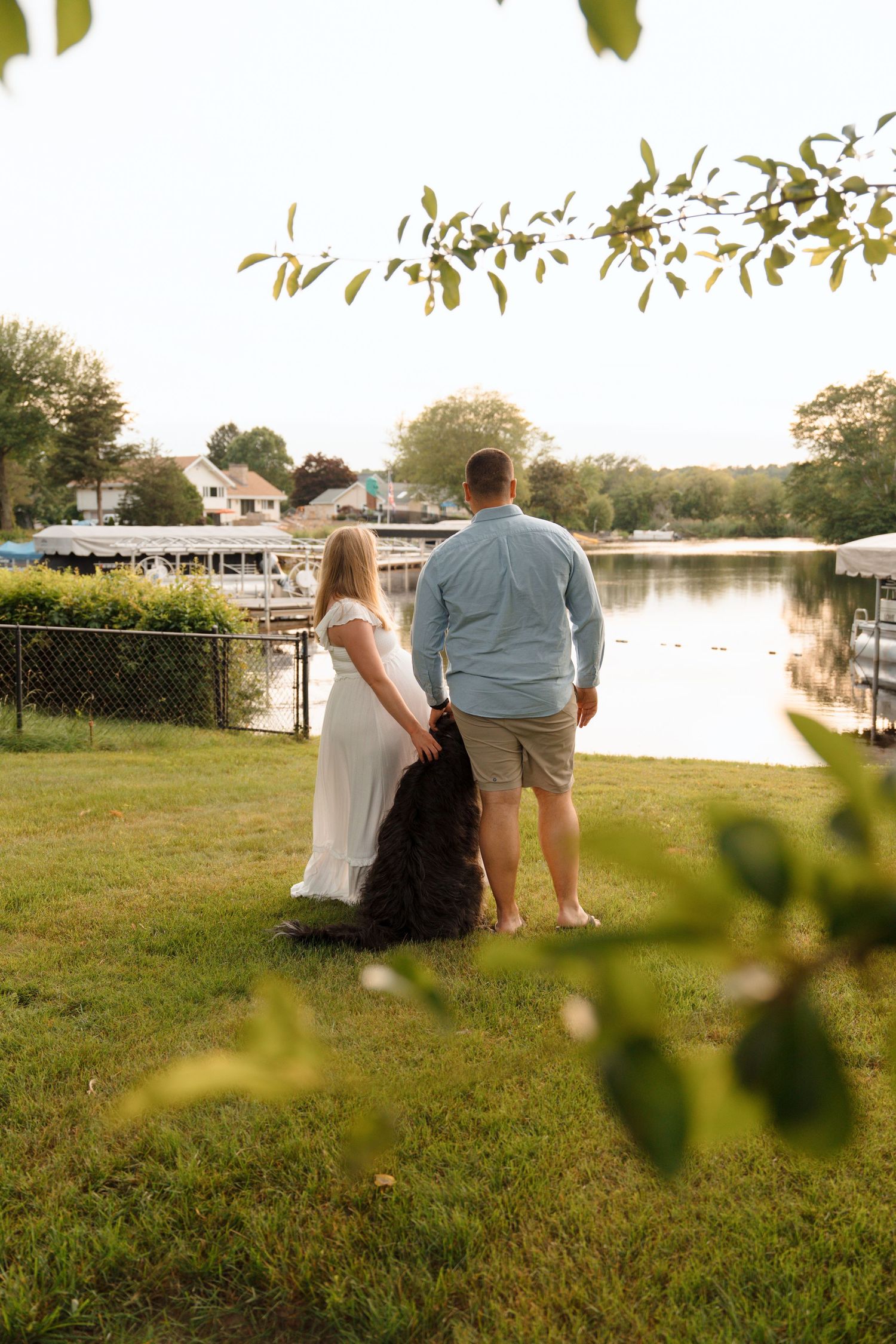 A couple sits together watching sunset on a grassy lawn by a calm lake or river.