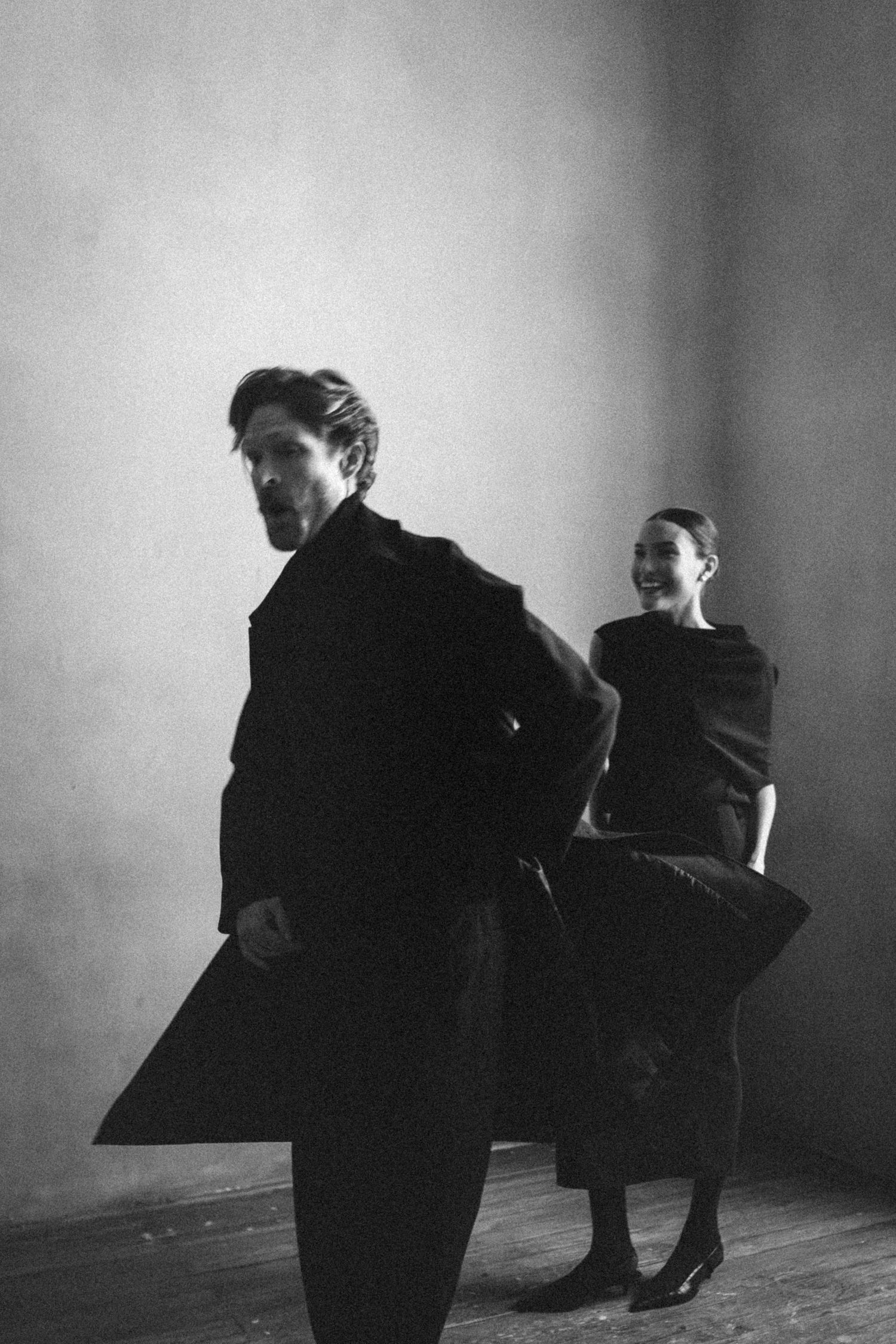 Black and white photo of man in dark coat and smiling woman in elegant black dress posing in minimalist room.