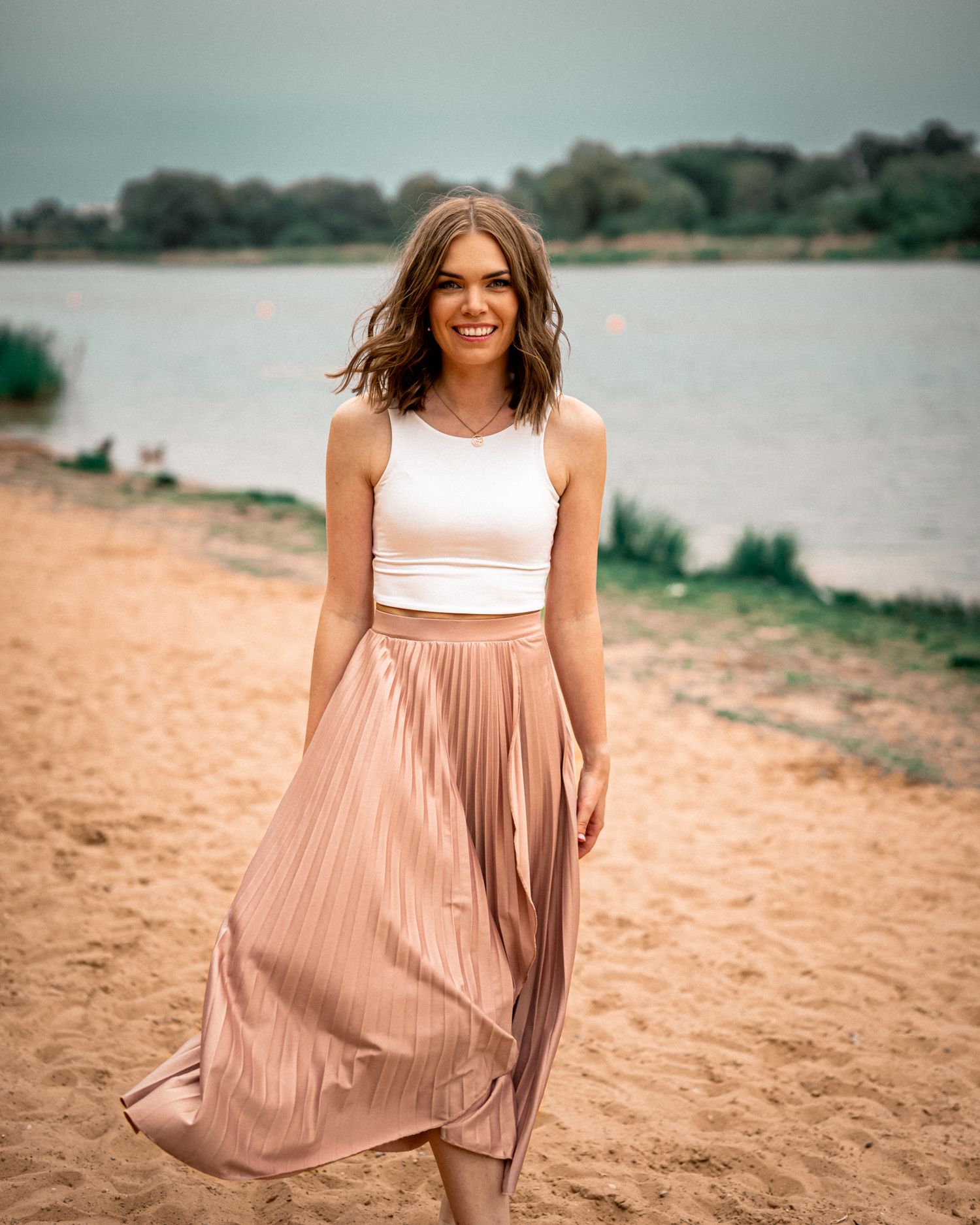 Smiling woman in white crop top and rose gold pleated midi skirt posing on sandy beach by lake.