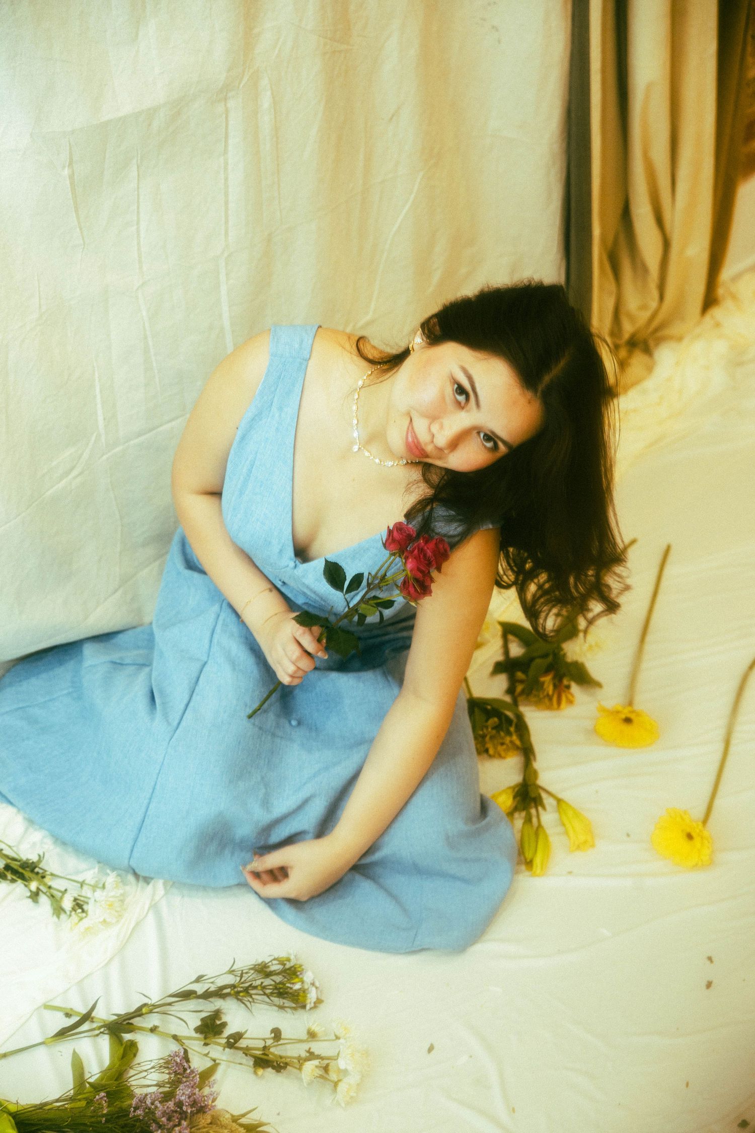 Young woman in blue dress sitting on floor holding red rose surrounded by scattered flowers near draped fabric backdrop.