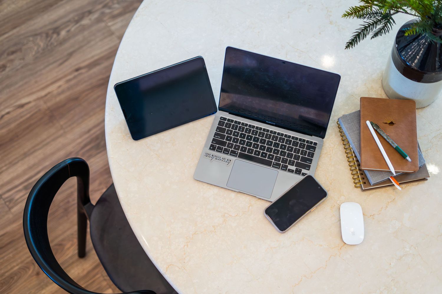 Overhead view of a modern workspace with a laptop, tablet, smartphone, mouse, notebooks, and a plant on a round white table.