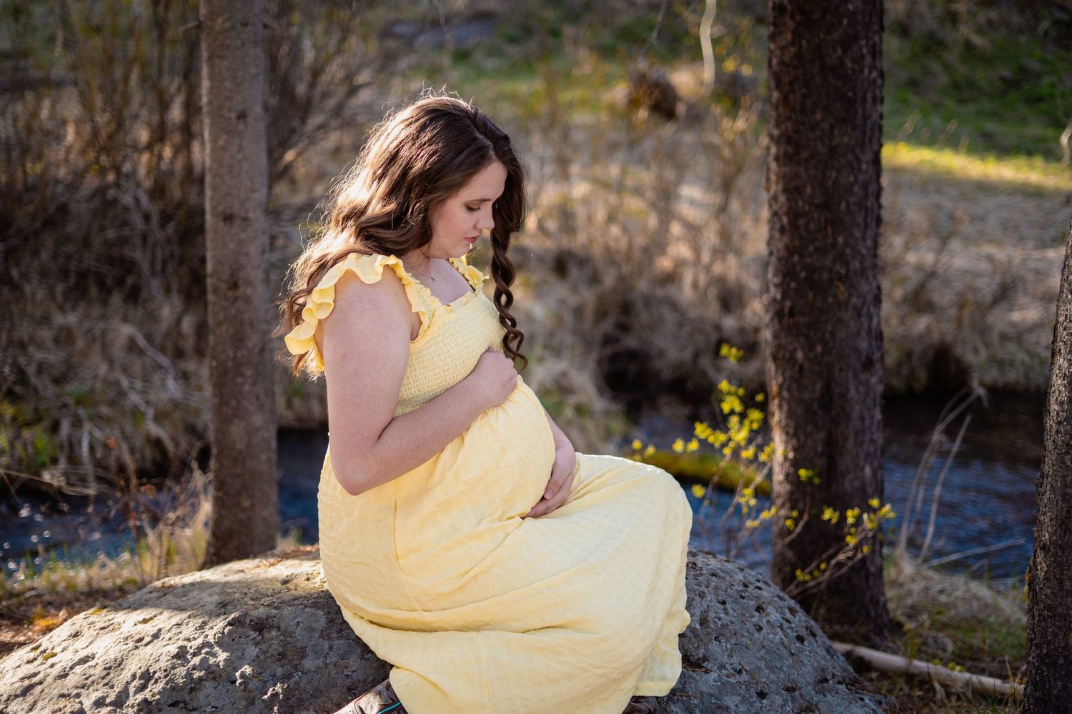 Pregnant woman in yellow dress sitting on a rock by a stream in golden light.
