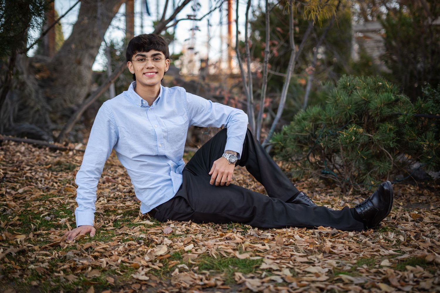 Young man in blue shirt sitting casually on fallen autumn leaves outdoors.