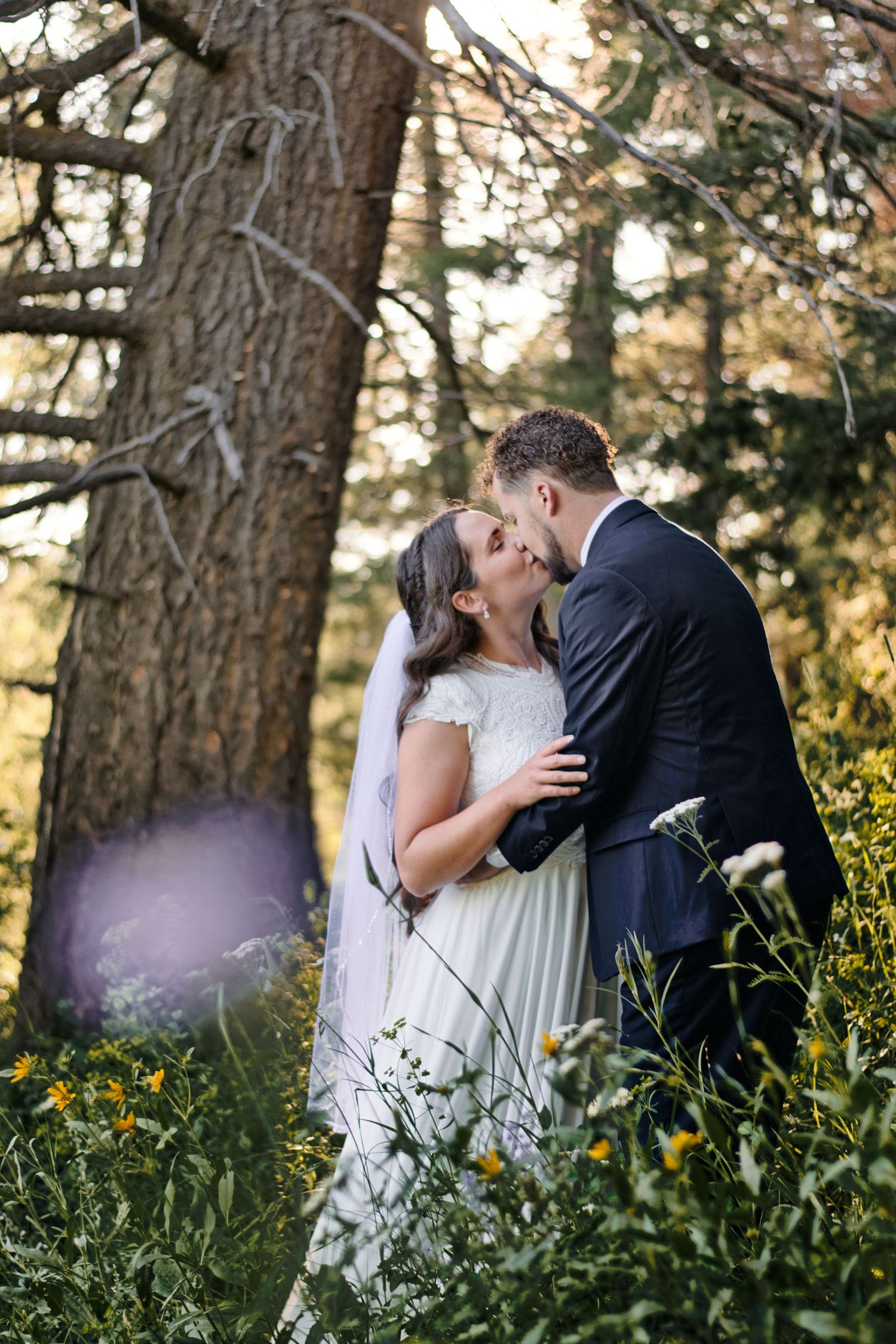 Bride and groom sharing a kiss among wildflowers beneath tall pine trees.