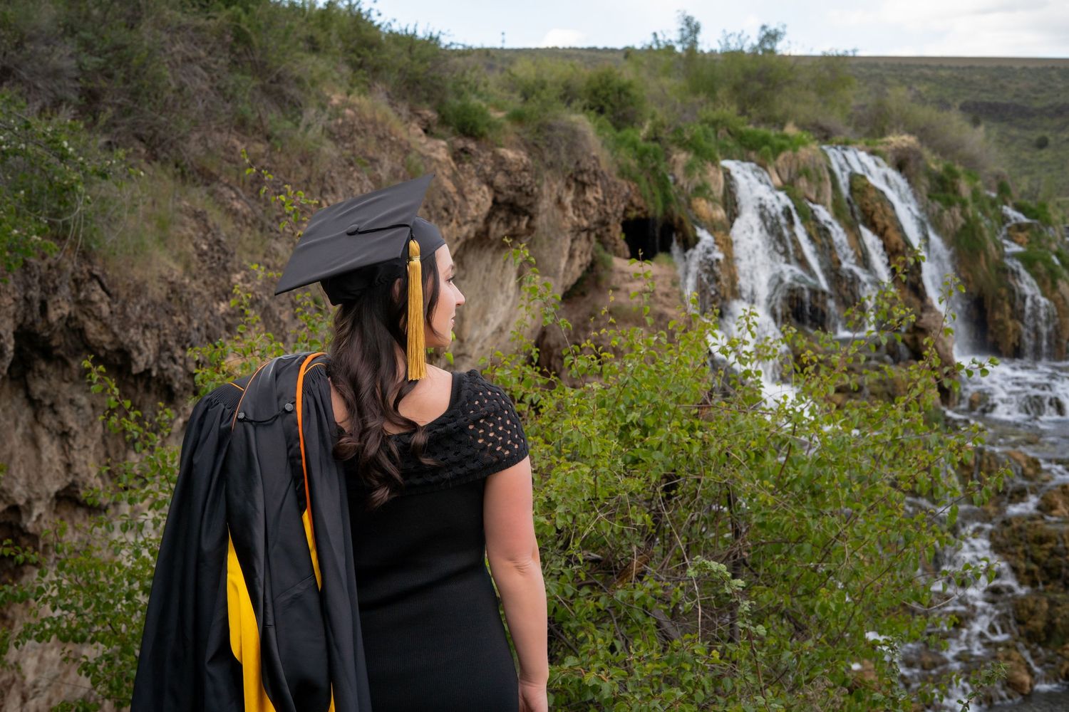 A graduate in cap and gown gazes at a scenic waterfall surrounded by lush greenery during an outdoor graduation photo session.