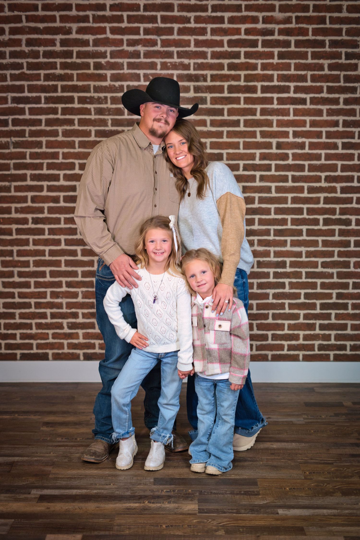 Family of four posing in front of a brick wall, father in black cowboy hat with two young daughters and mother.