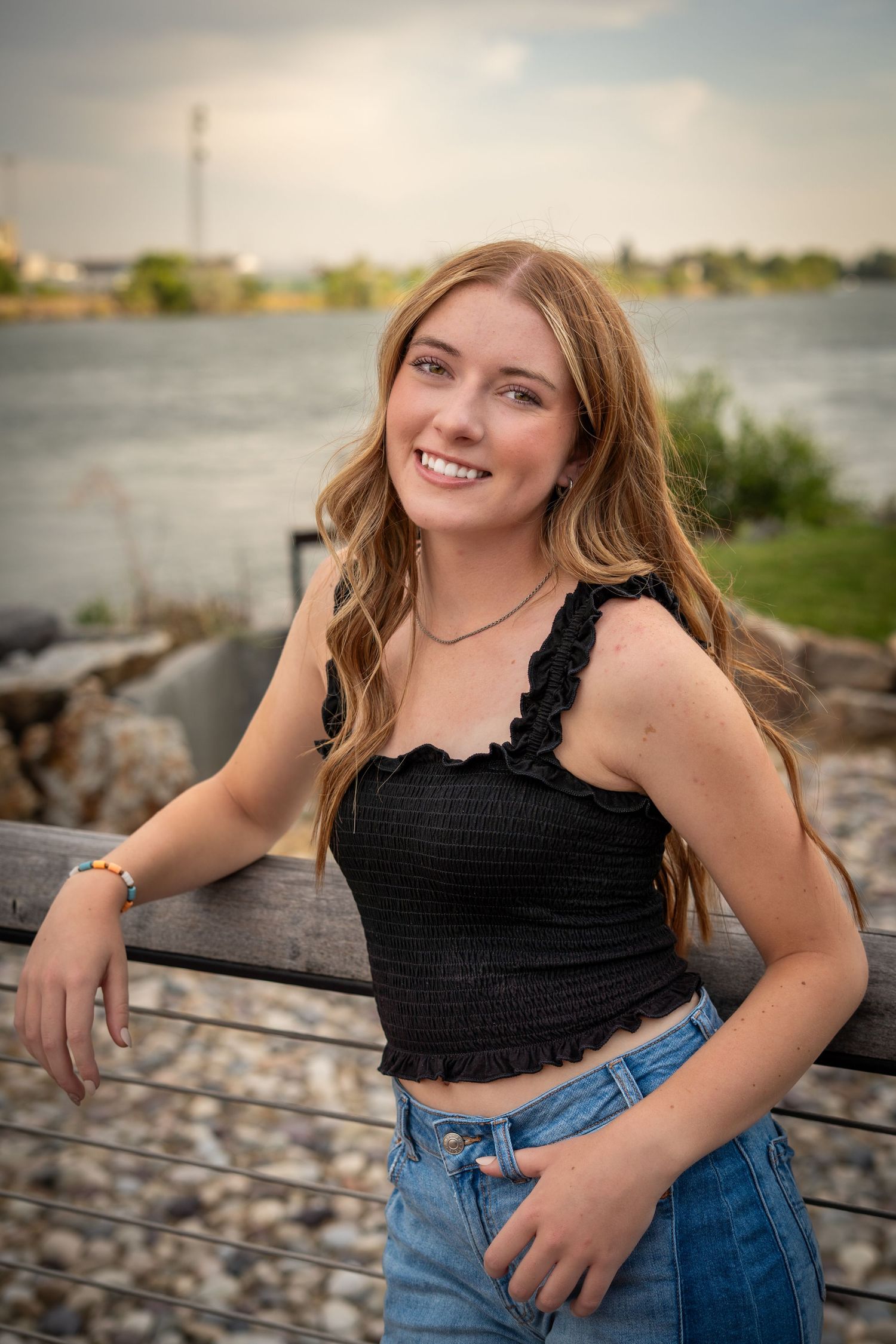Smiling teenage girl in black crop top and jeans leans on railing with waterfront view behind her.