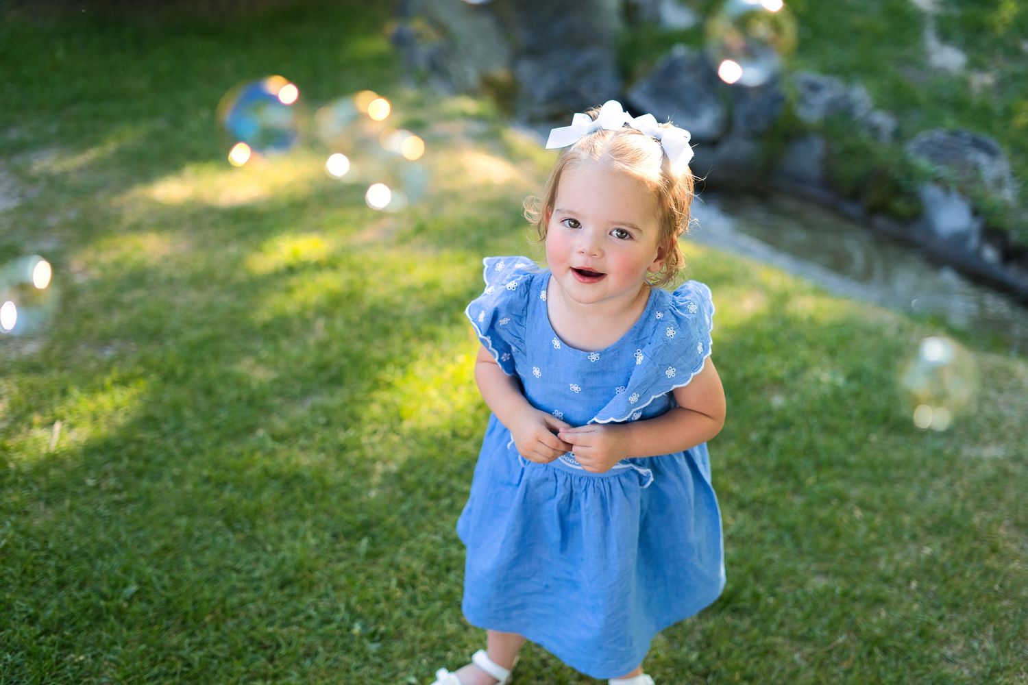 Adorable toddler girl in blue polka dot dress playing with bubbles on green grass outdoors.