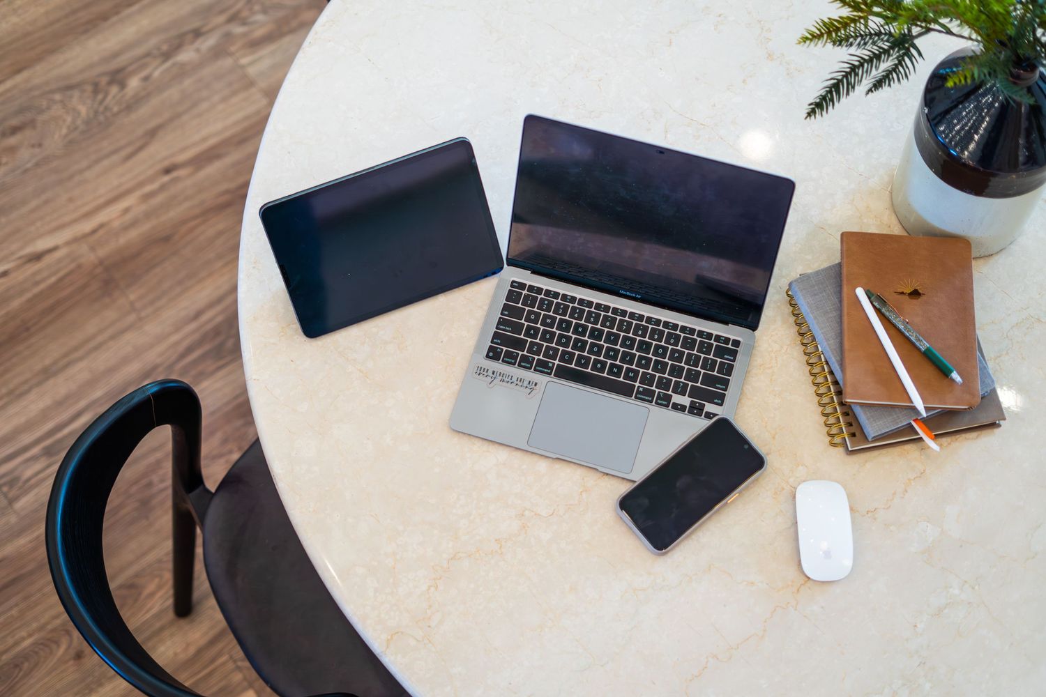 Overhead view of a laptop, tablet, smartphone, mouse, notebooks, and plant on a round white table.