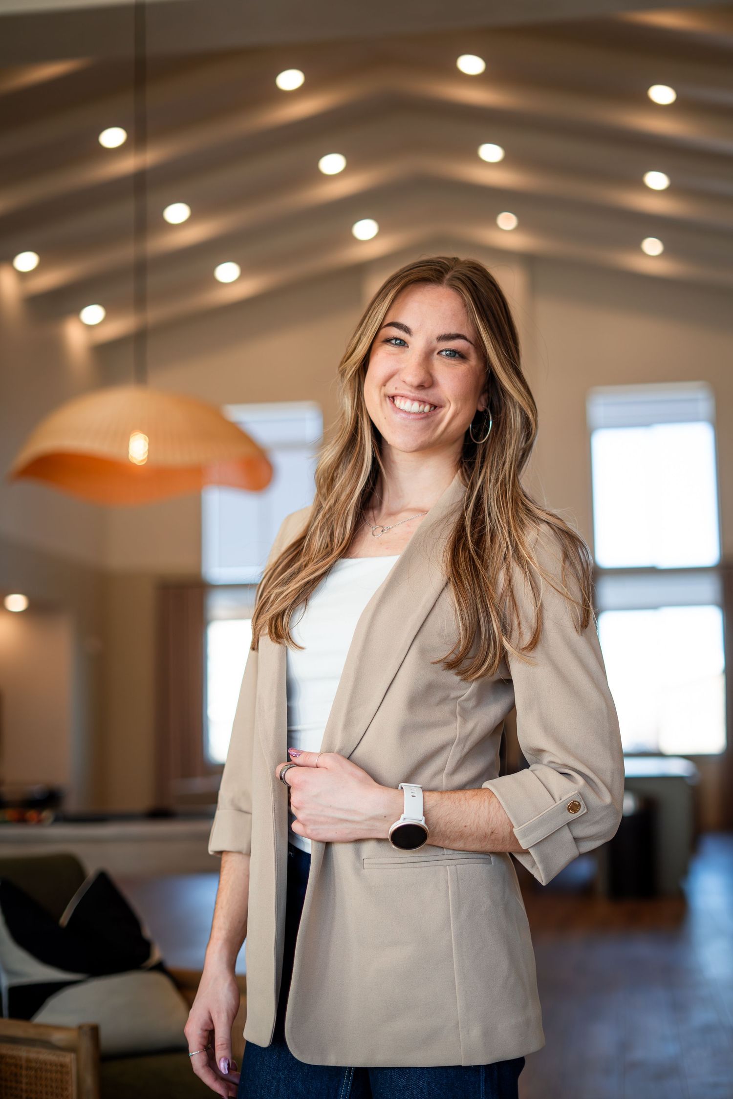 Smiling young professional woman in beige blazer standing in modern office with vaulted ceiling.