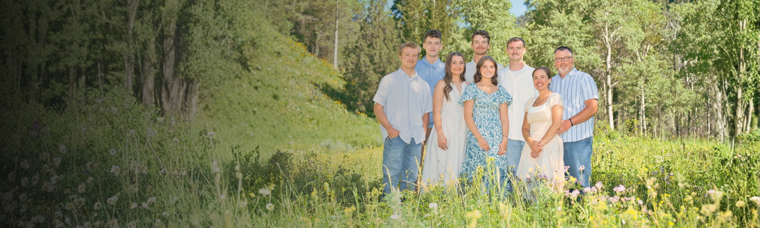 A happy family of seven poses outdoors in a lush green meadow surrounded by tall trees on a sunny day.