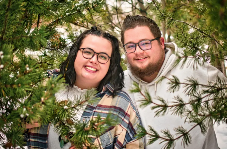 Smiling couple with glasses posing together among snow-dusted pine tree branches.
