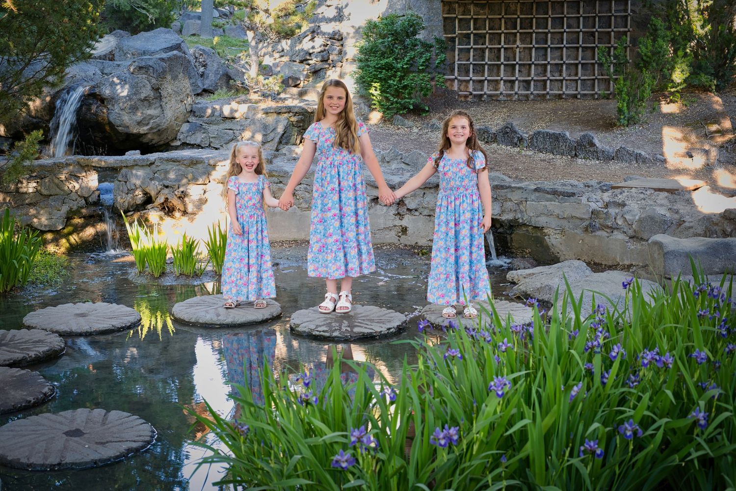 Three girls in matching floral dresses holding hands while standing on stepping stones over a garden pond.