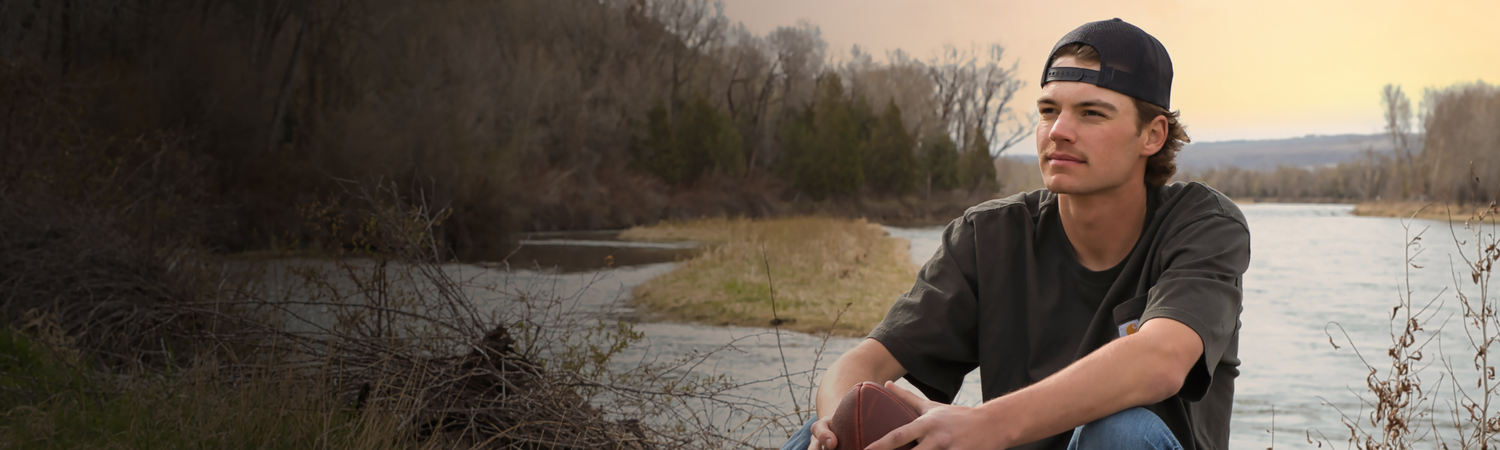 Young man in baseball cap sitting by a calm river with bare trees in golden sunset light.