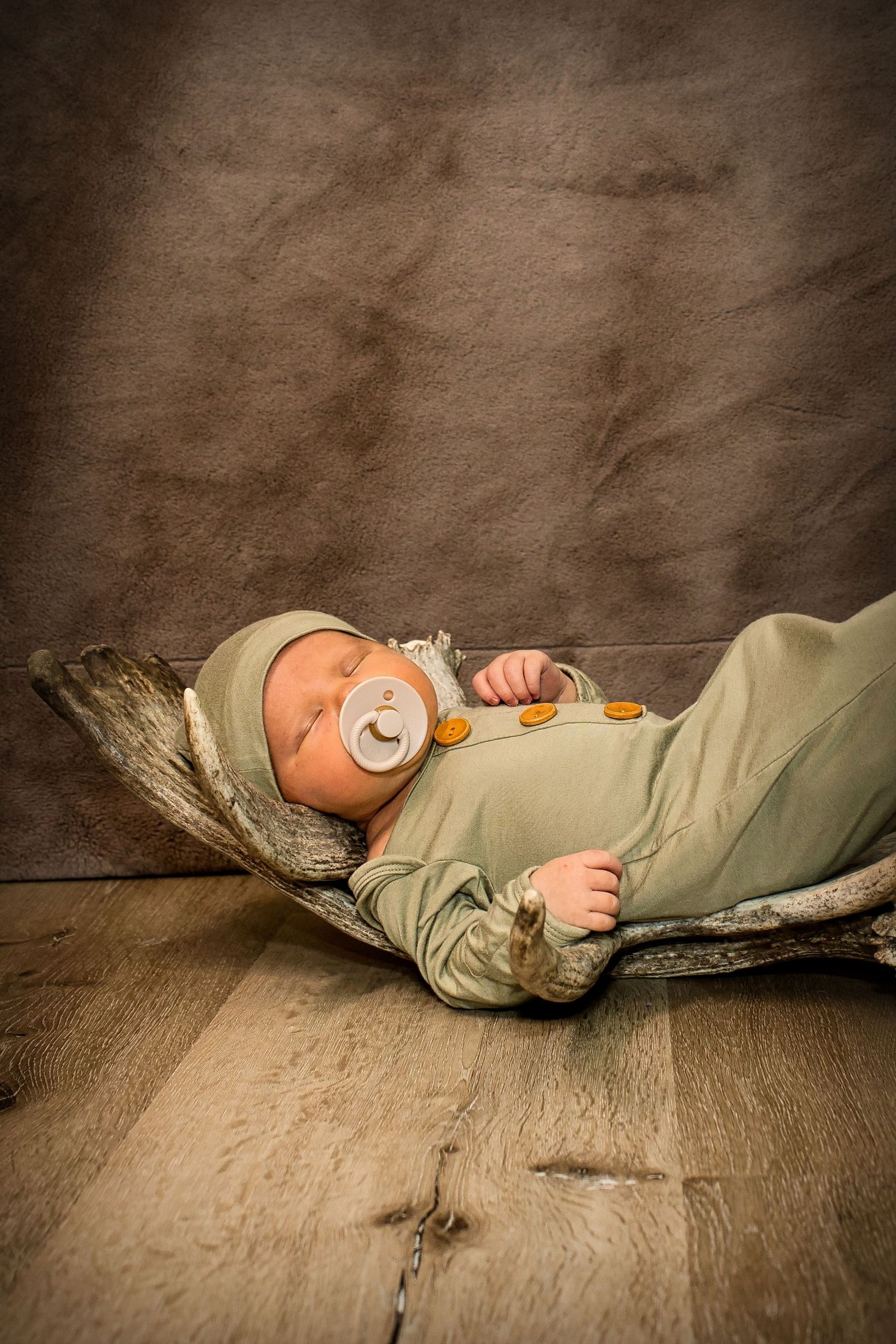 Newborn baby sleeping peacefully in a rustic wooden cradle wearing a green outfit and hat with a pacifier.