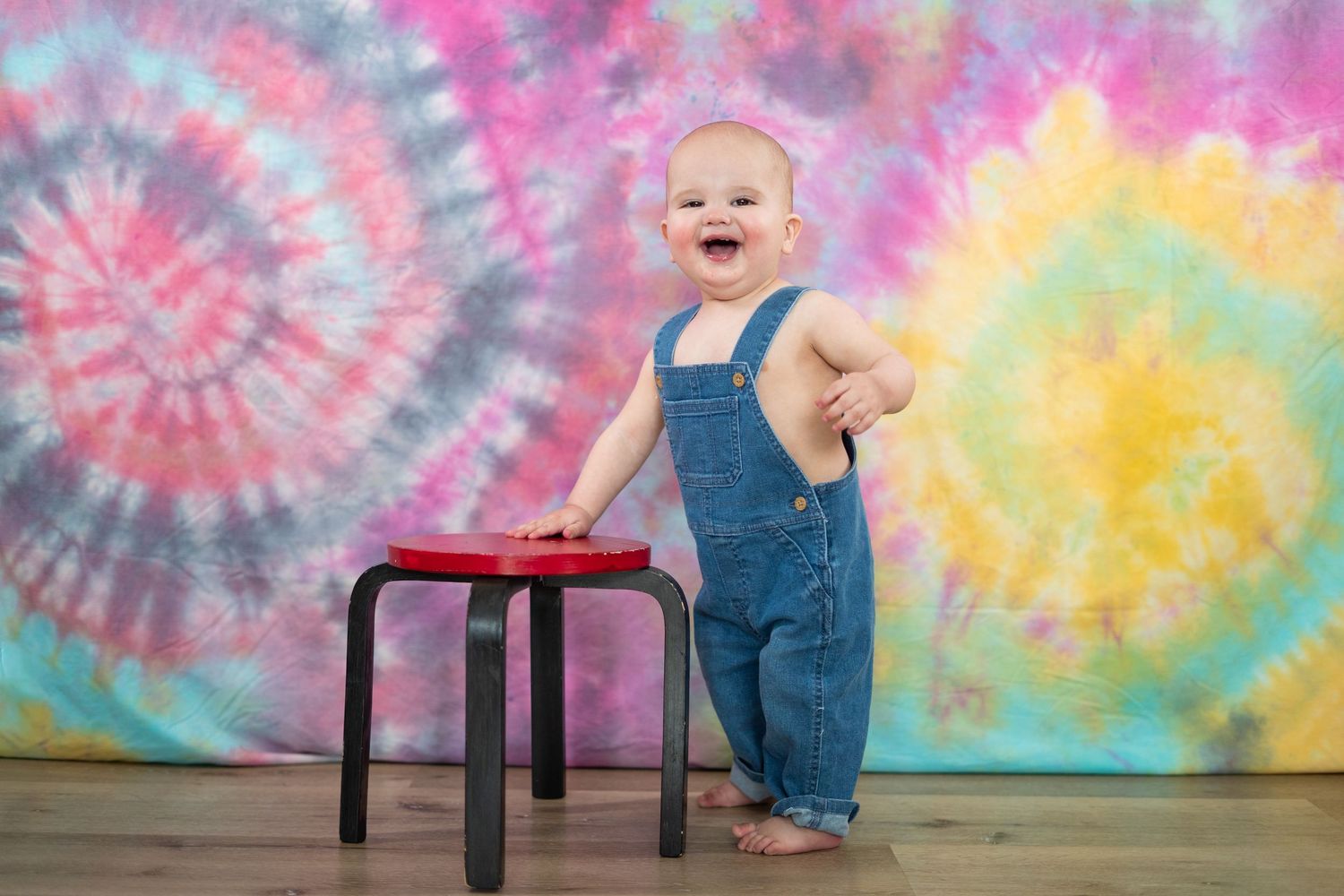 Happy toddler in denim overalls laughs while standing at small table against colorful tie-dye backdrop.