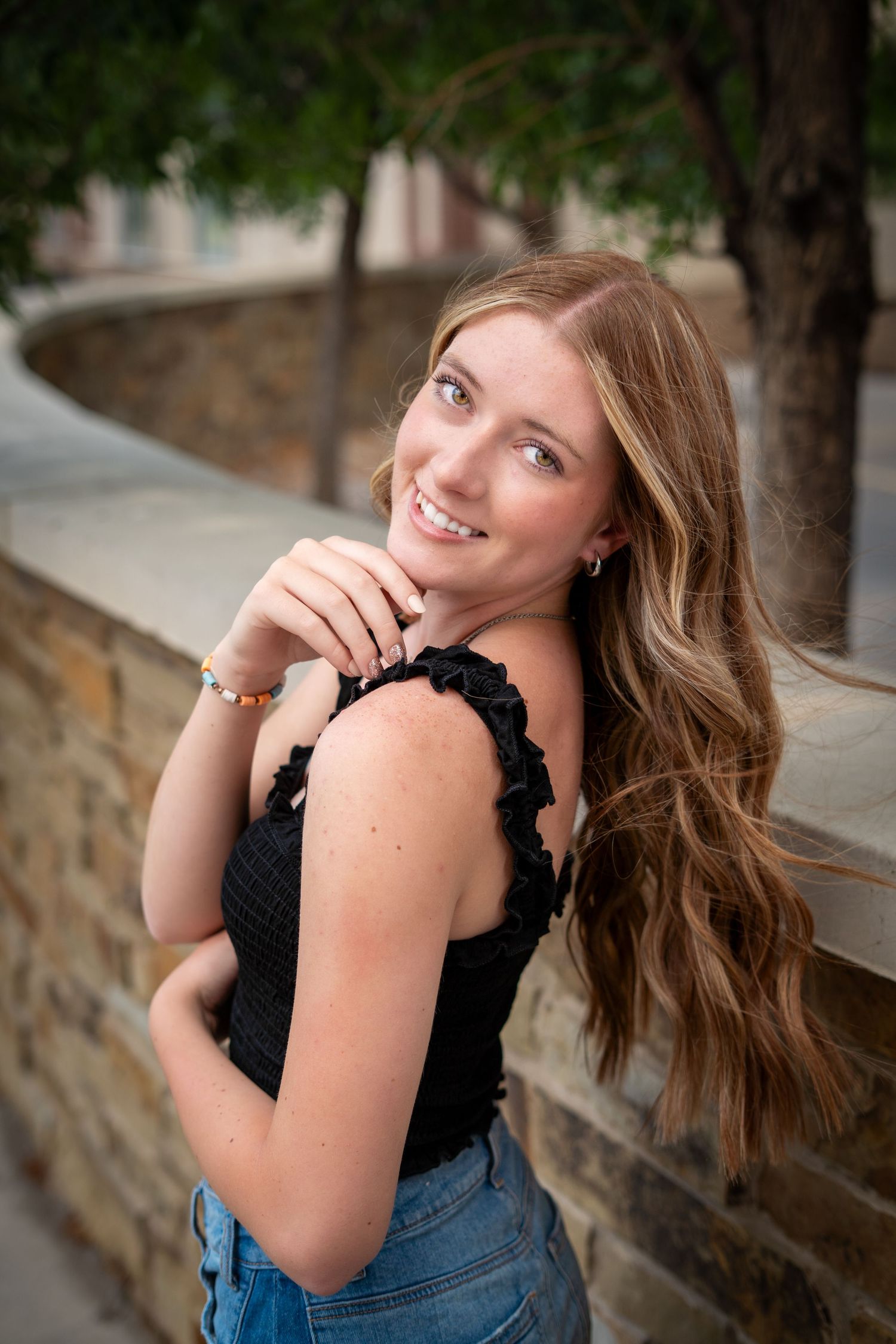 Smiling teenage girl with long wavy hair in black top and jeans poses by a stone wall outdoors.