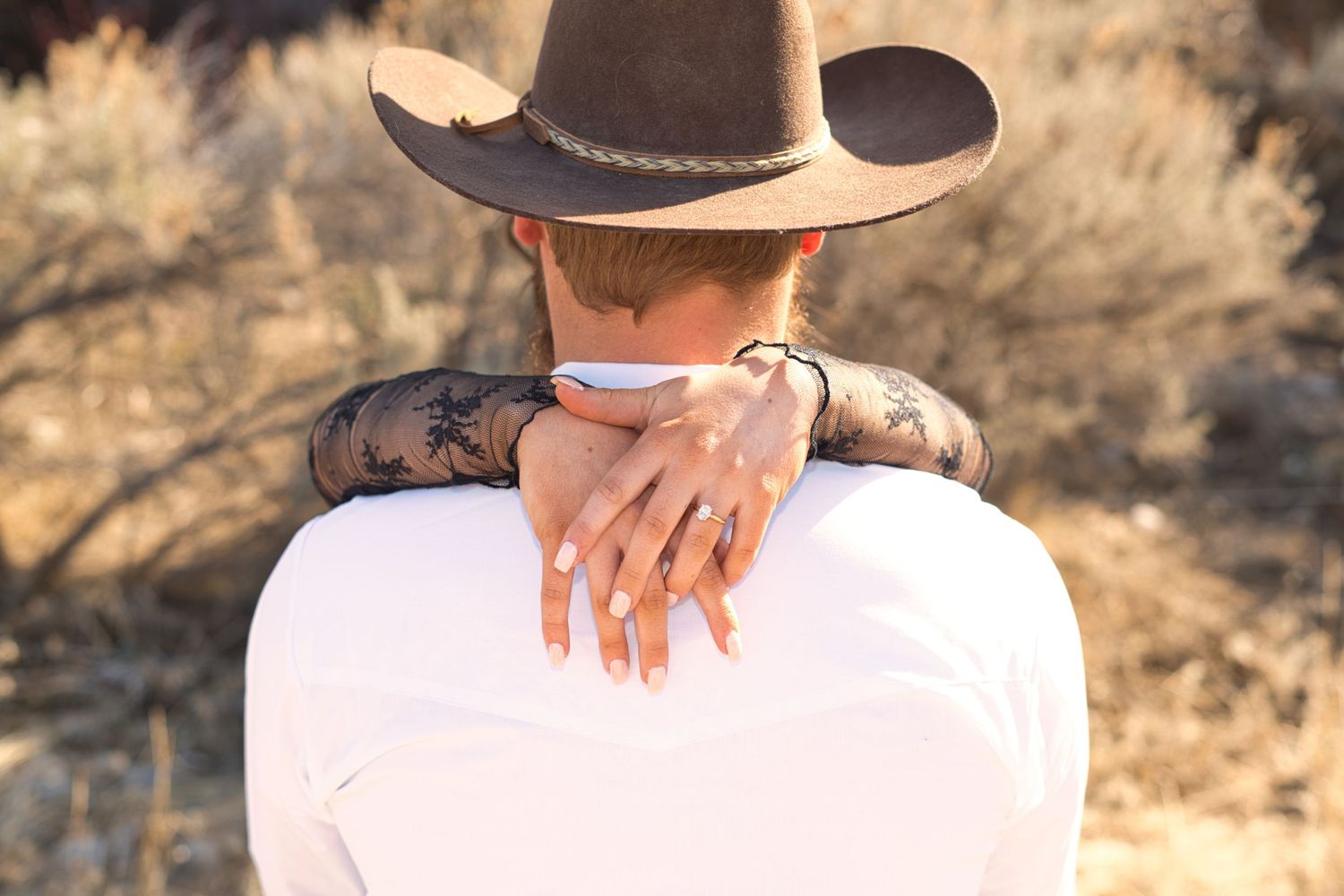 Couple embracing outdoors, woman wearing a cowboy hat with a tattoo visible on her arm, man in white shirt.