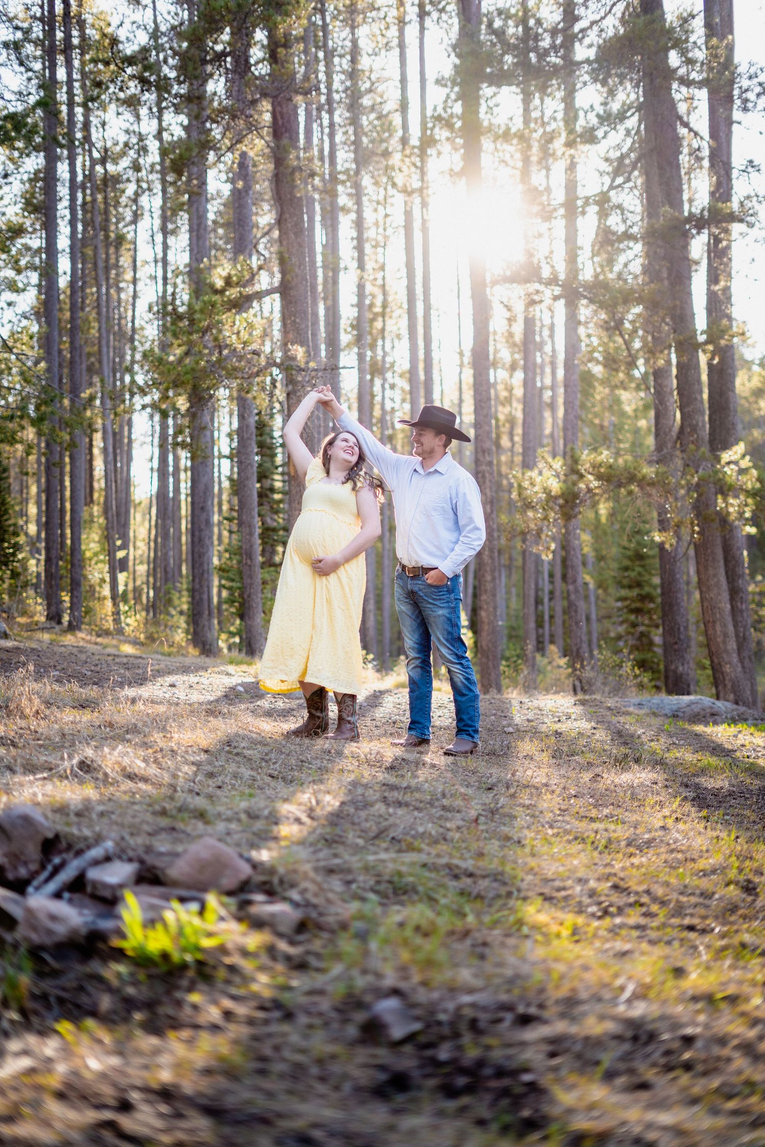 Expecting couple dancing outdoors on a sunlit forest path, woman in yellow maternity dress and man in cowboy hat.