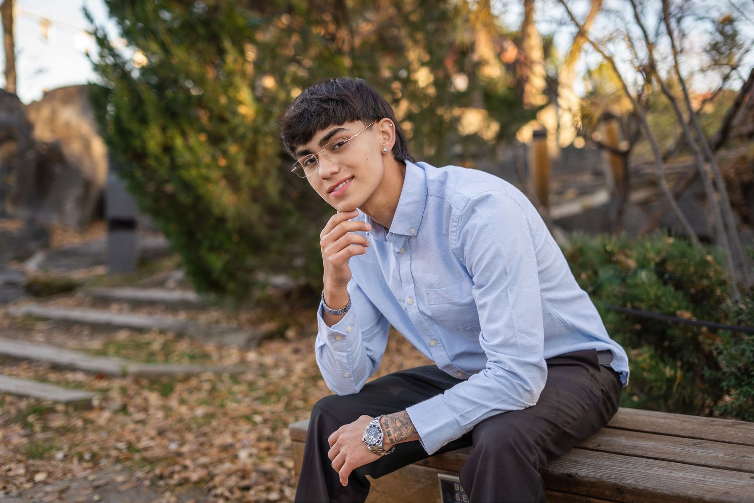 Teen boy in light blue shirt sits on bench smiling in an outdoor park setting with fall foliage.