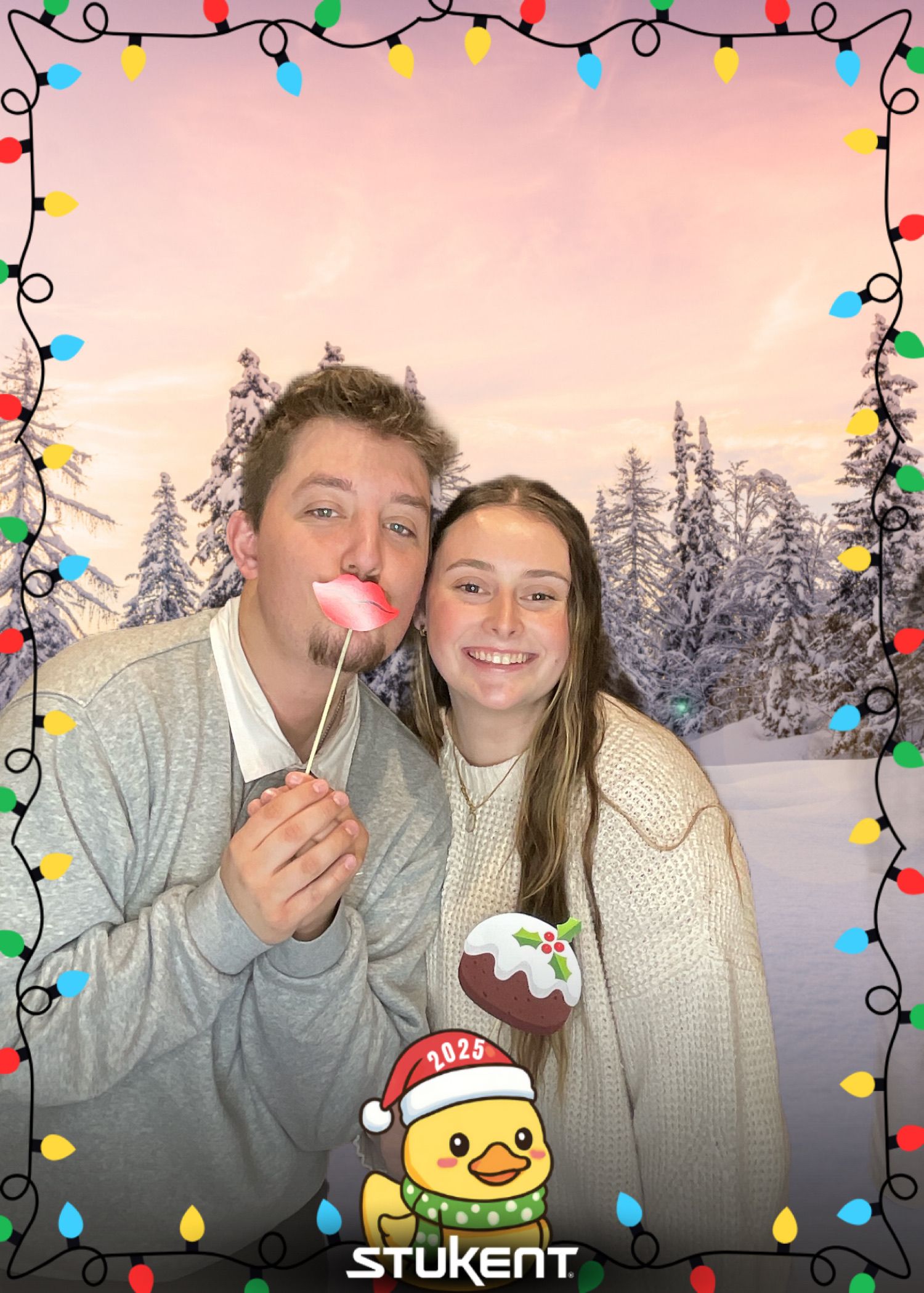 Two teens pose for a festive Christmas photo booth picture with holiday lights border and snowy winter forest background.