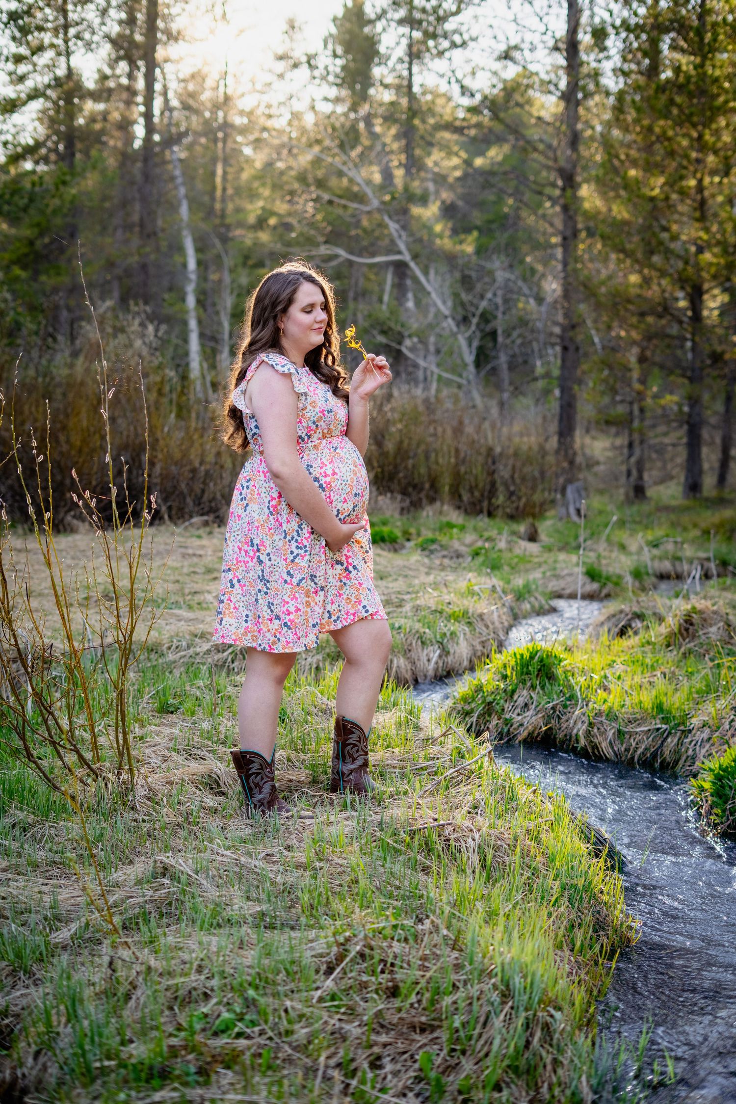Pregnant woman in floral dress and cowboy boots standing by a stream in a sunlit forest, smelling a flower.