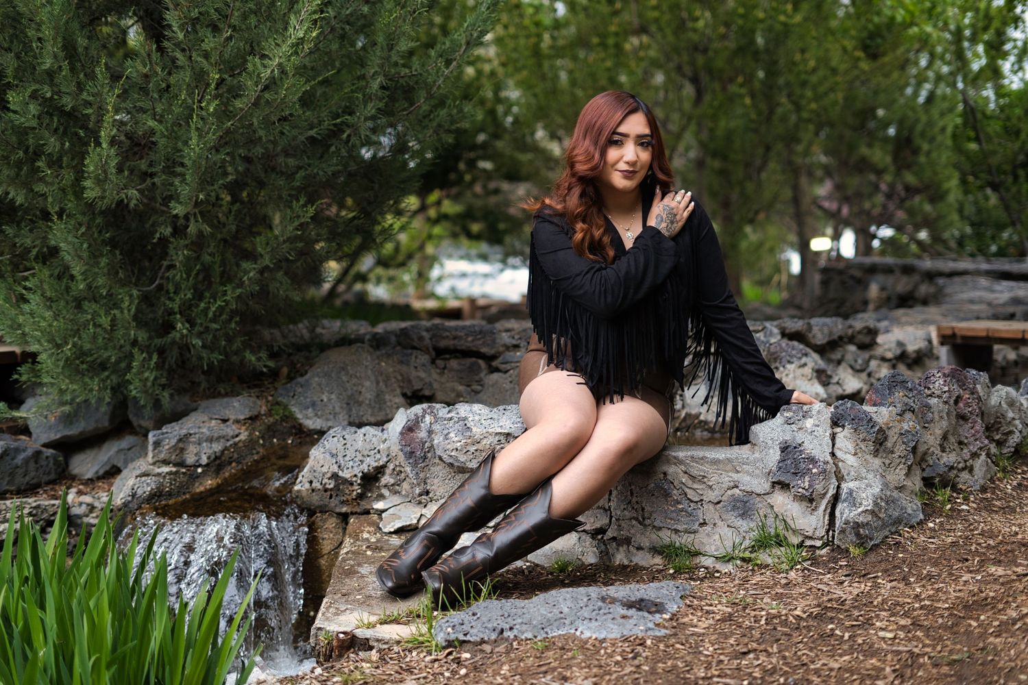 Young woman in black fringe top and knee-high boots sitting on rocks in a lush garden.