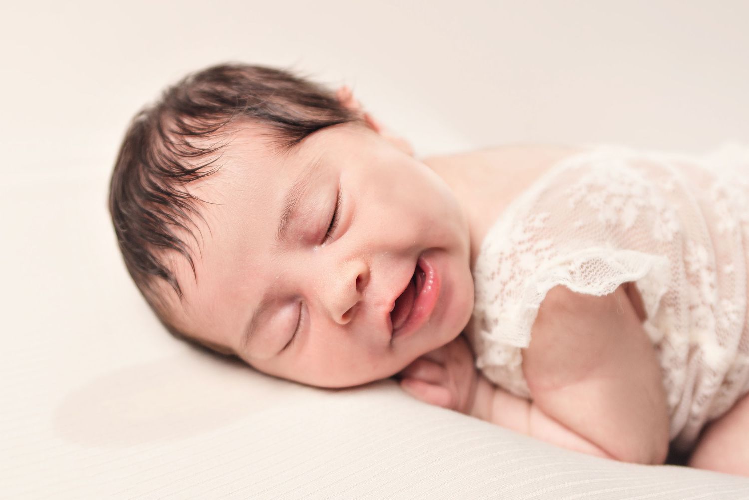 A peaceful sleeping baby wearing white lace clothing lies on a soft white blanket in a serene pose.