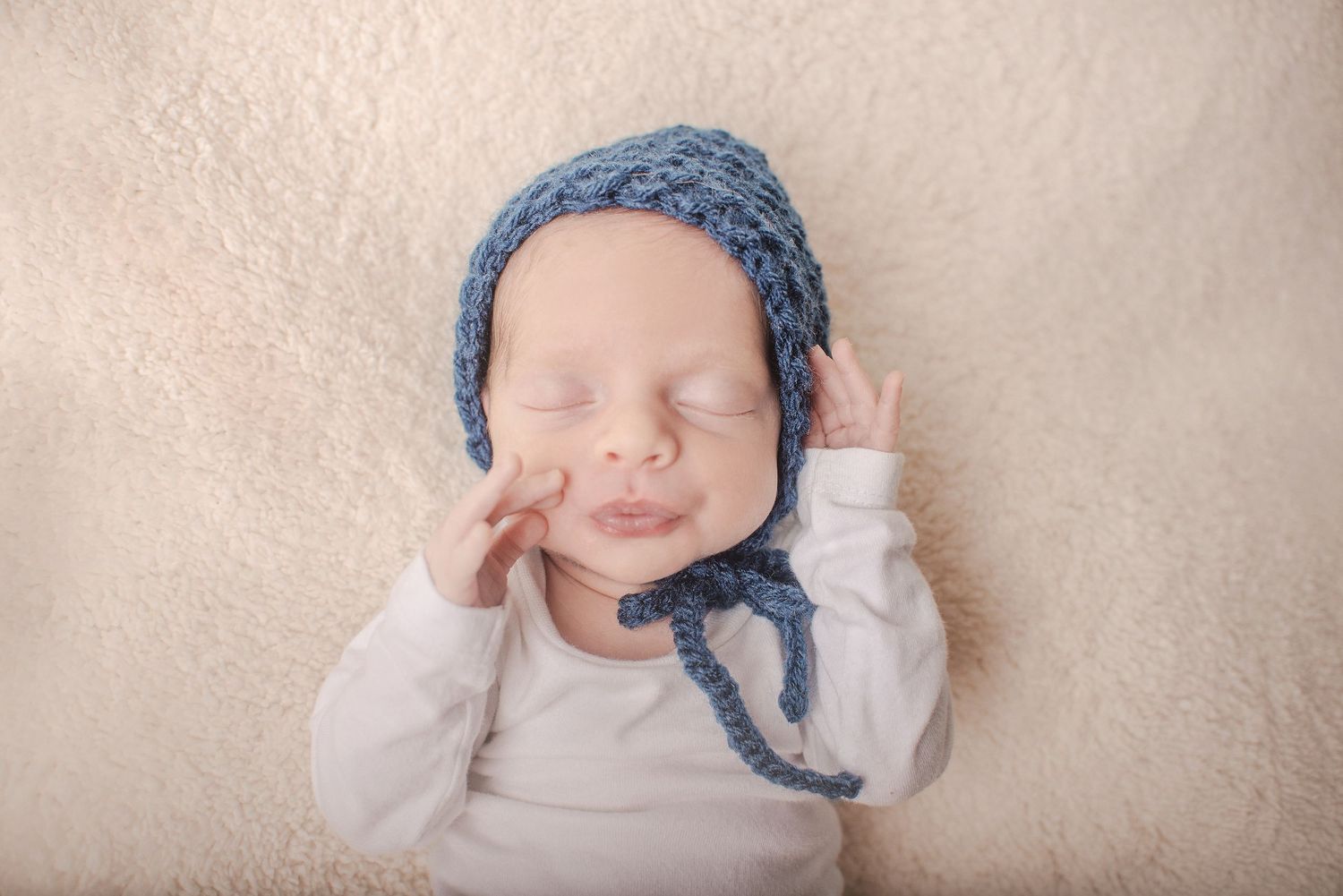 Sleeping newborn wearing blue knit bonnet on soft white backdrop.