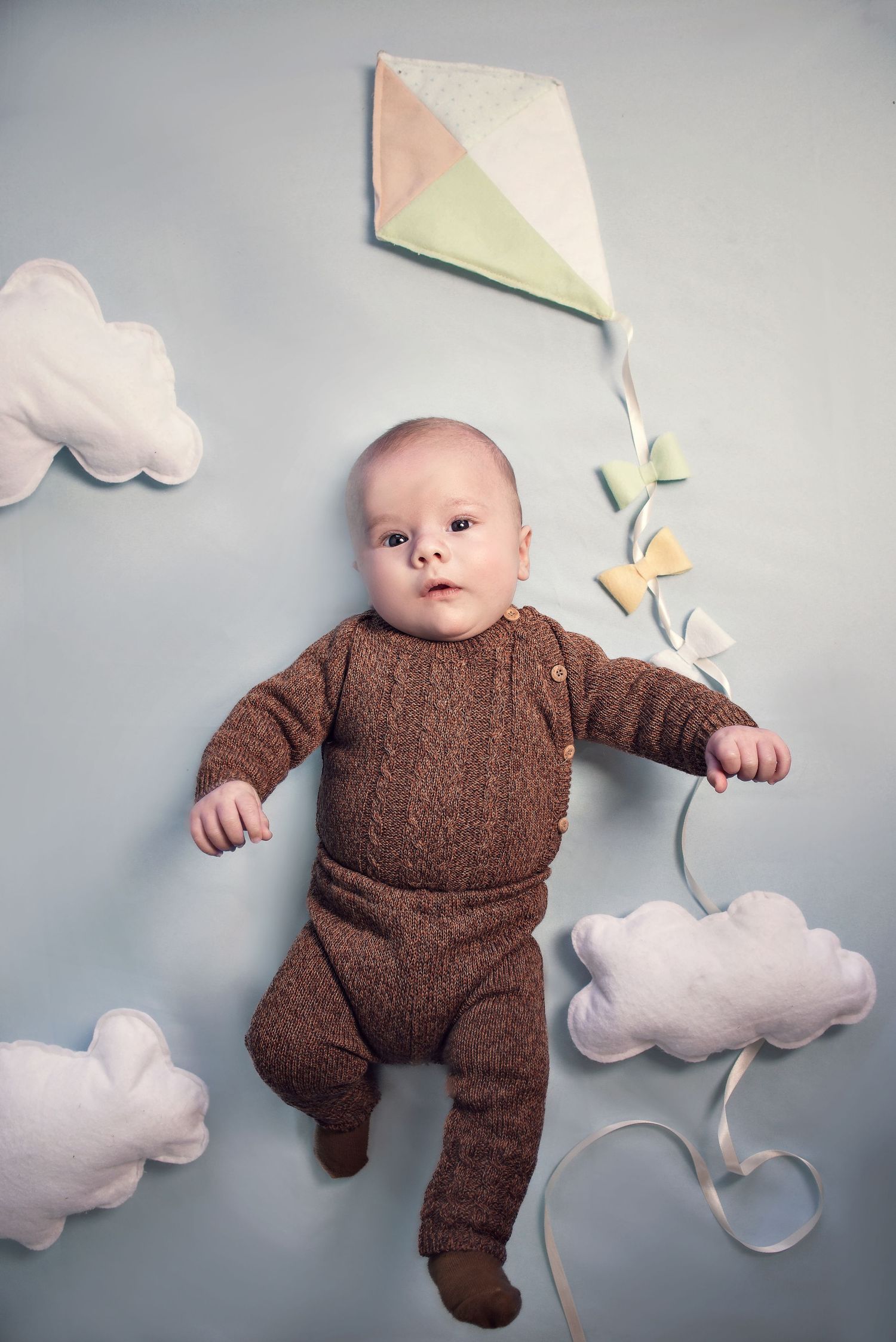 Baby in brown knit outfit lying on gray background with decorative white paper clouds and butterflies.