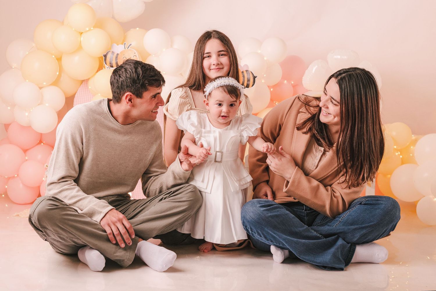 Family wearing neutral colored clothing poses together on floor with soft pink and peach balloons in background.