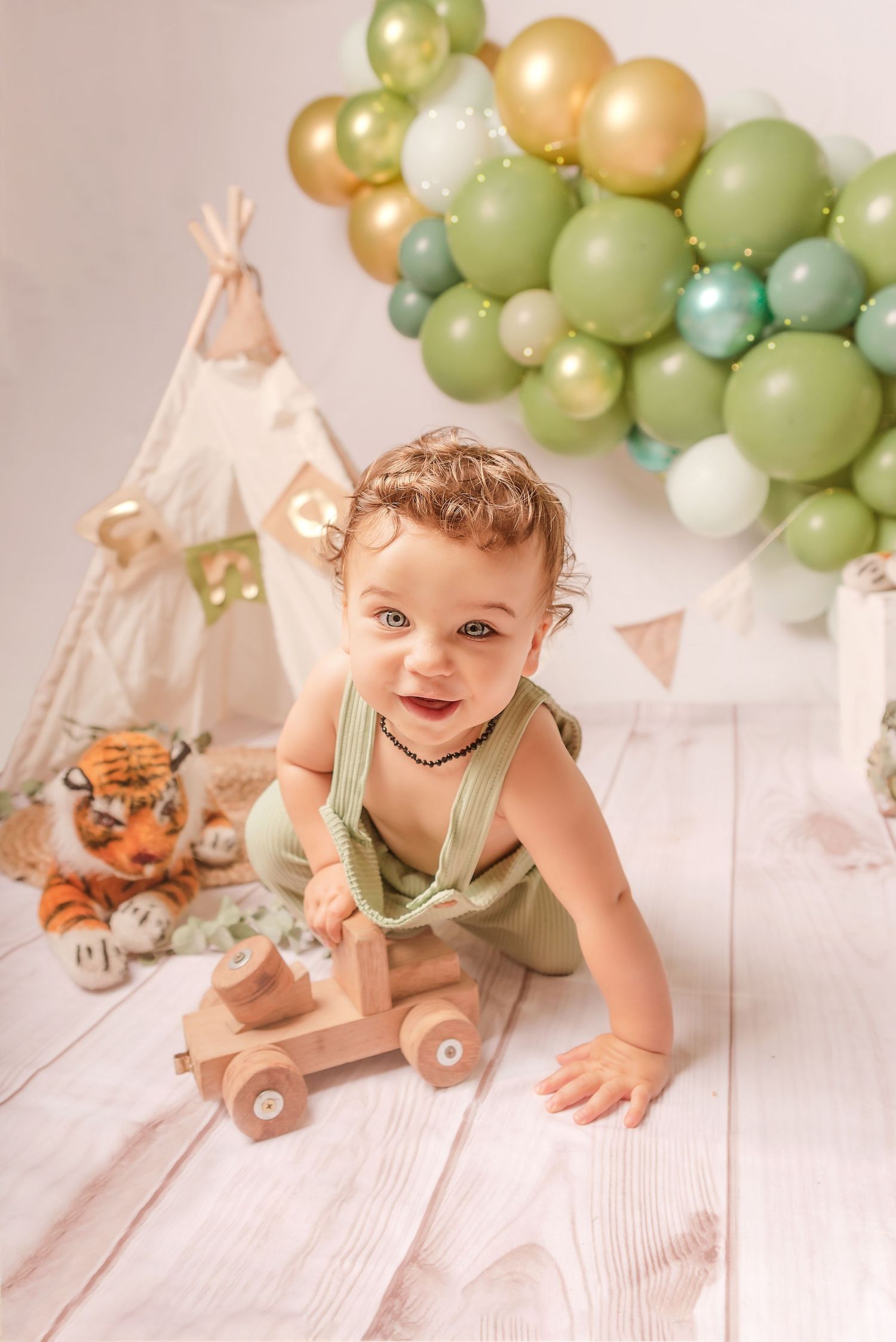 Balloon arch in soft green and gold tones decorates a white backdrop with small teepee decorations.