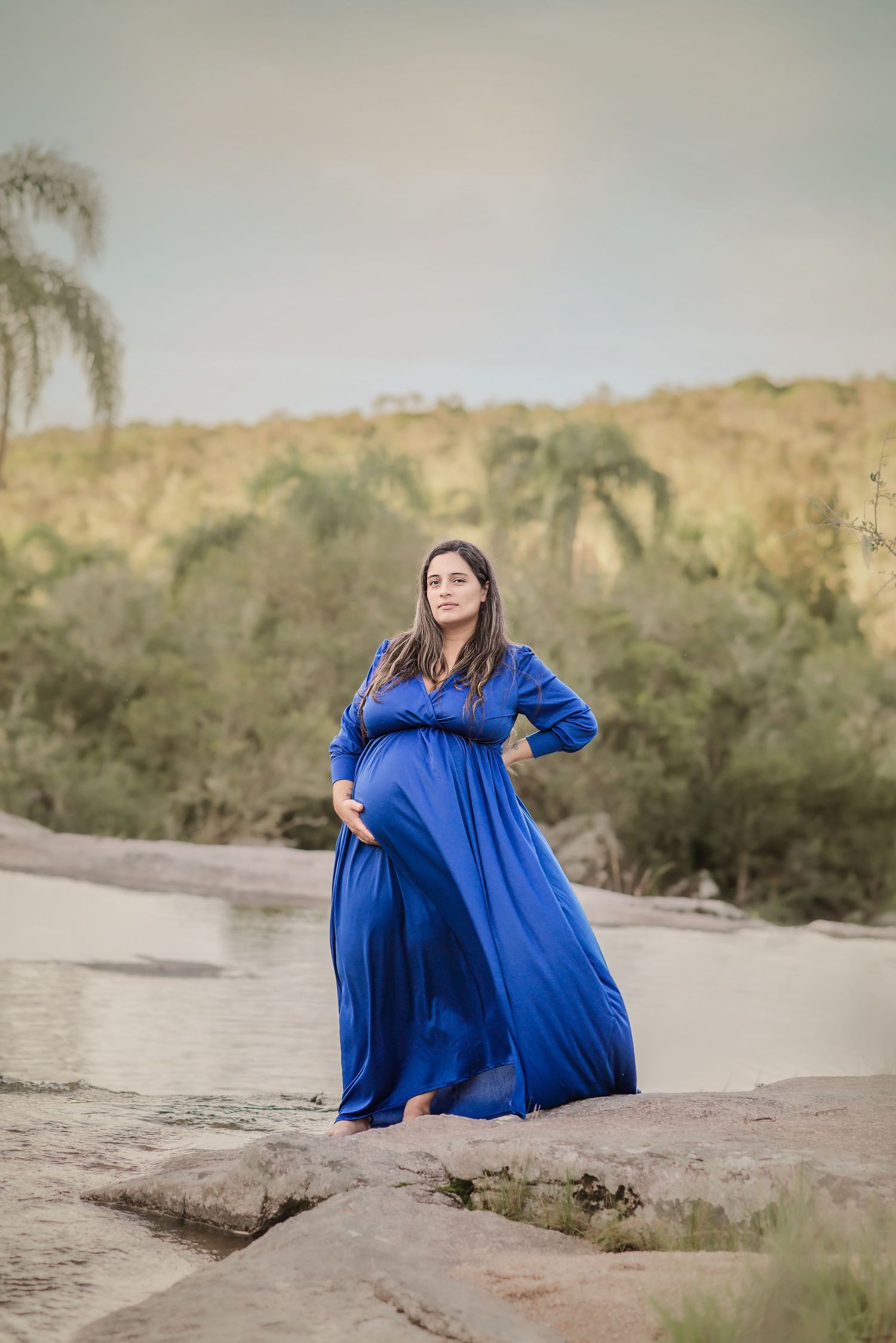 A woman in a flowing royal blue dress poses gracefully in a desert landscape with palm trees and hills in the background.
