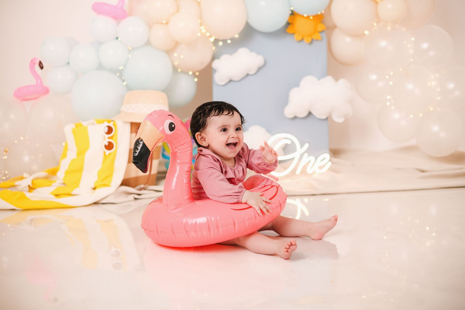 A dreamy baby photo session setup with soft pink decorations, white clouds, and whimsical elements against a light backdrop.