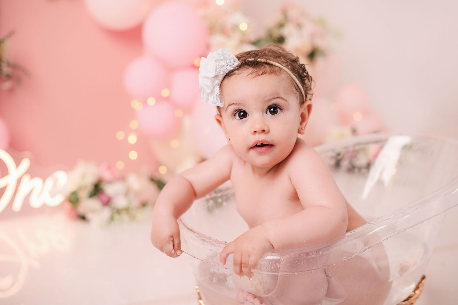 Baby in pink tutu dress sits on floor with sparkly pink bokeh light effects and floral decorations in background.