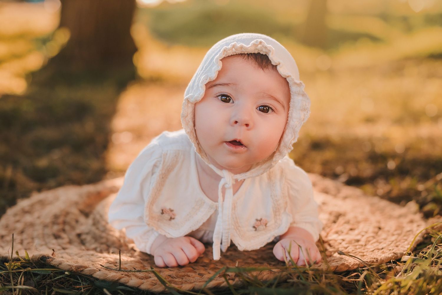 Baby in white bonnet and dress lying on autumn leaves in soft golden sunlight.