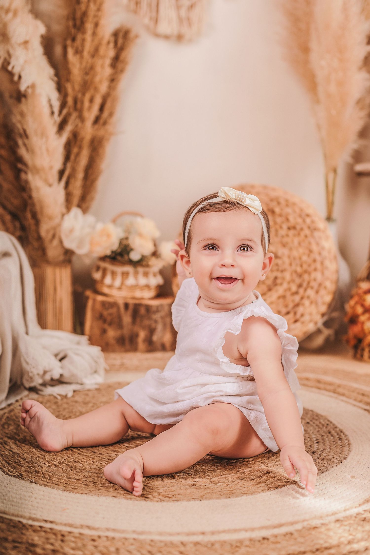 Baby in white dress sits on round woven rug surrounded by pampas grass and boho decor in warm neutral tones.