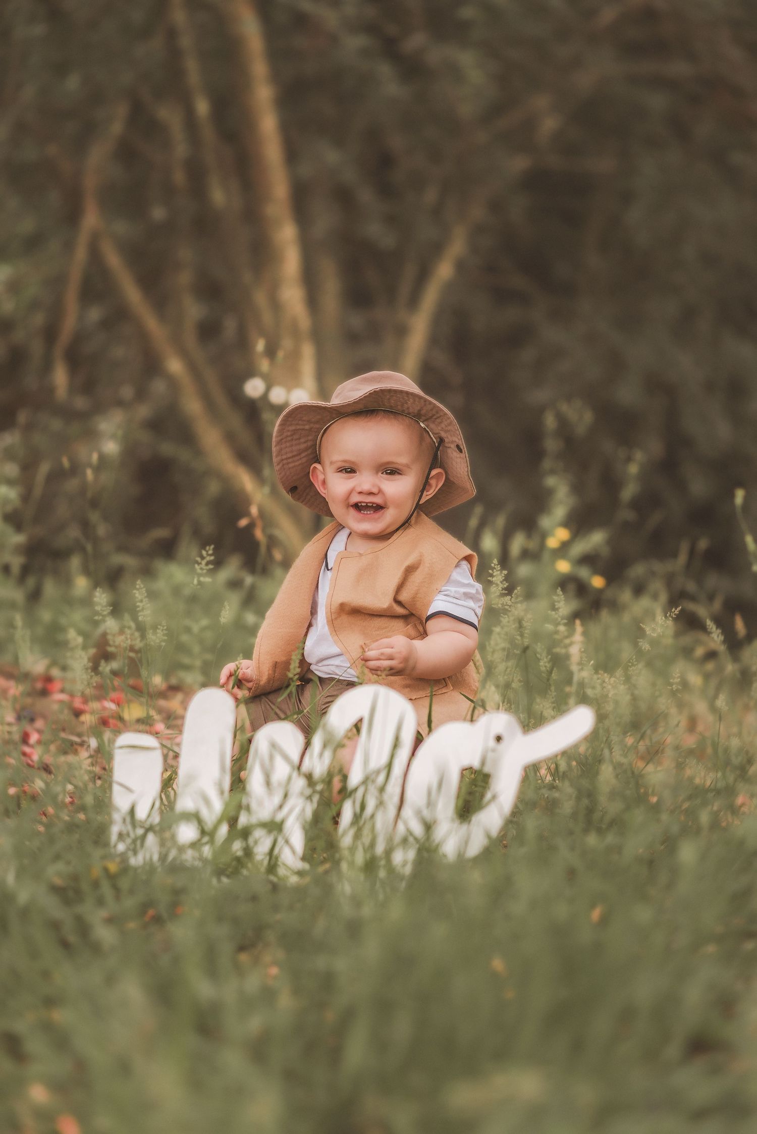 A baby in a brown hat sits in tall grass surrounded by white wooden letters in a natural outdoor setting.