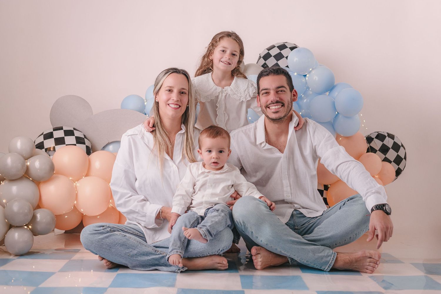 A happy family wearing matching denim and white outfits sits together on a light colored floor with peach balloons.
