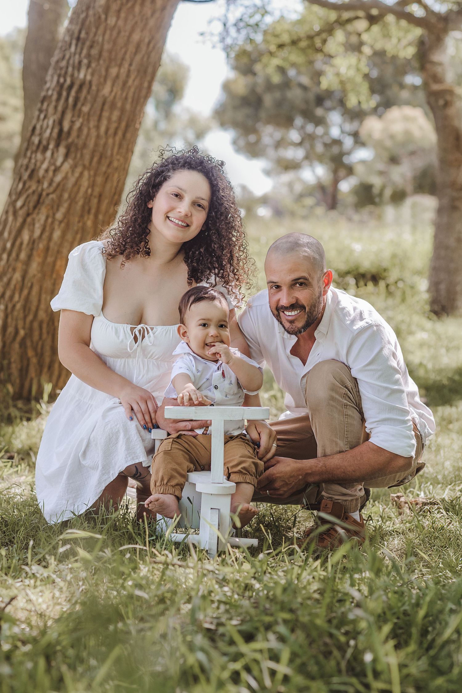 A family sits together on grass beneath tall trees on a sunny day, sharing joyful moments in matching white outfits.