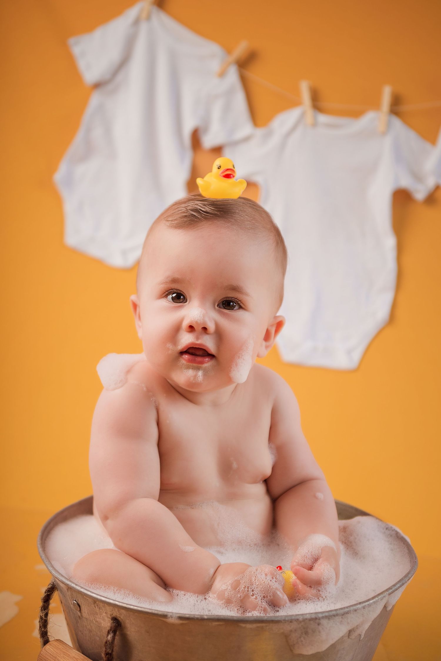 Baby bath time photo session with rubber duck and bubbles against vibrant orange background.