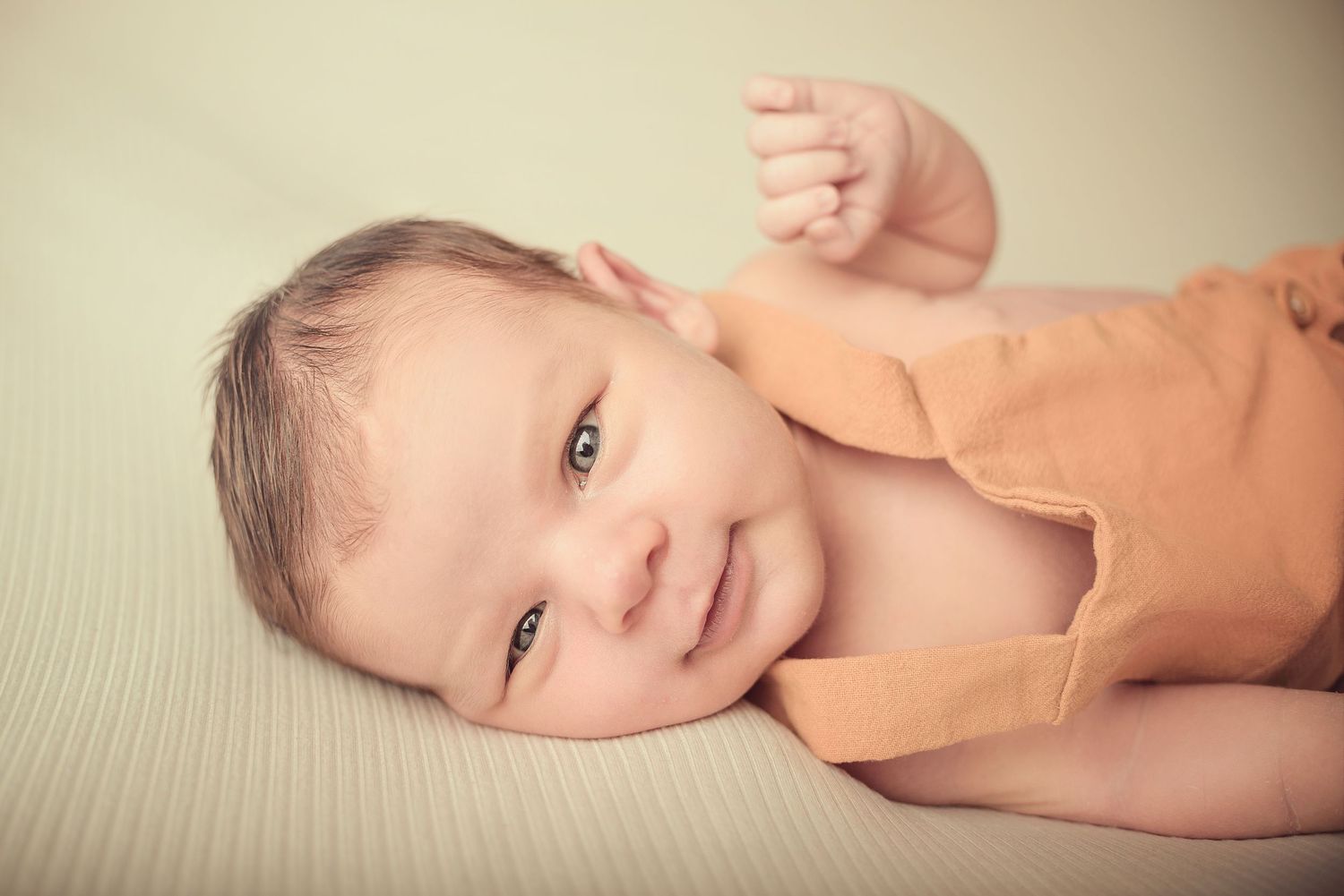 A smiling baby lies on a beige blanket with arms raised playfully in a warm-toned portrait.