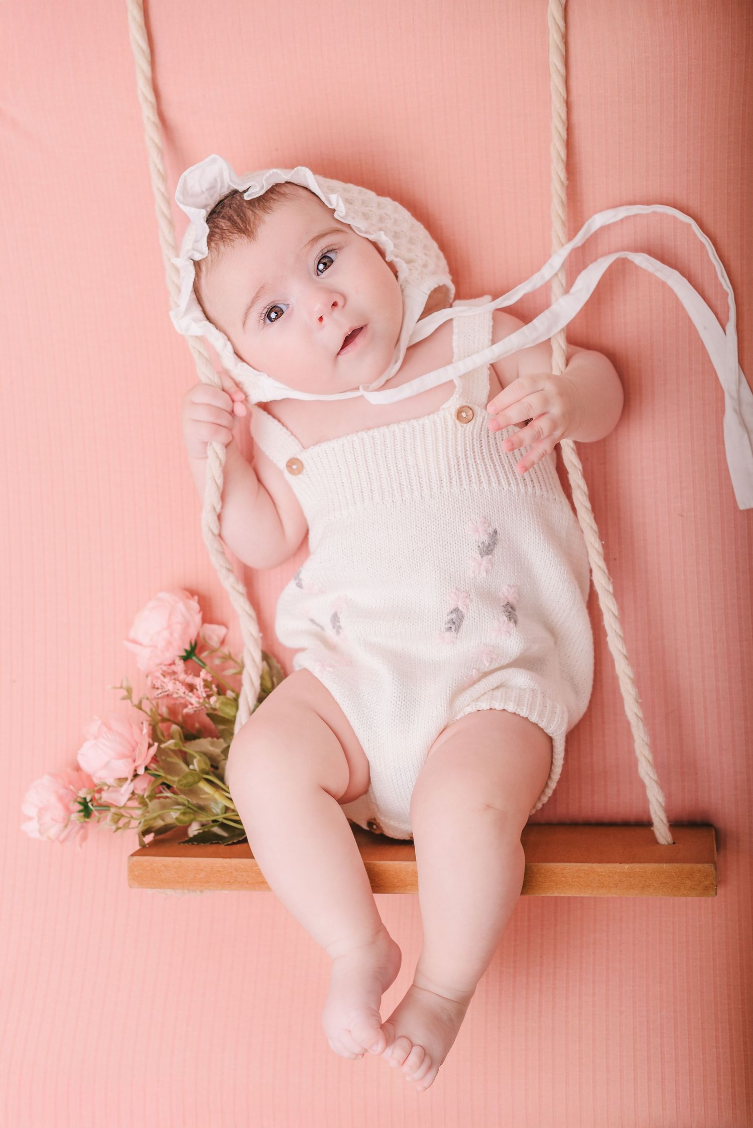 A baby in white clothing lies on a wooden swing against a pink background with delicate flowers.