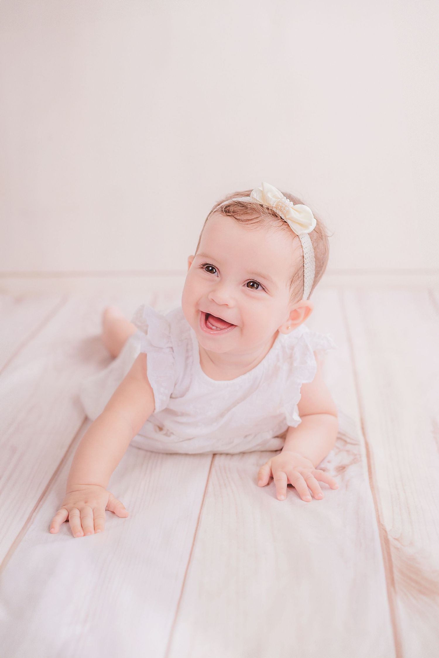 A baby wearing a white dress and bow headband lies on white bedding while smiling at the camera.