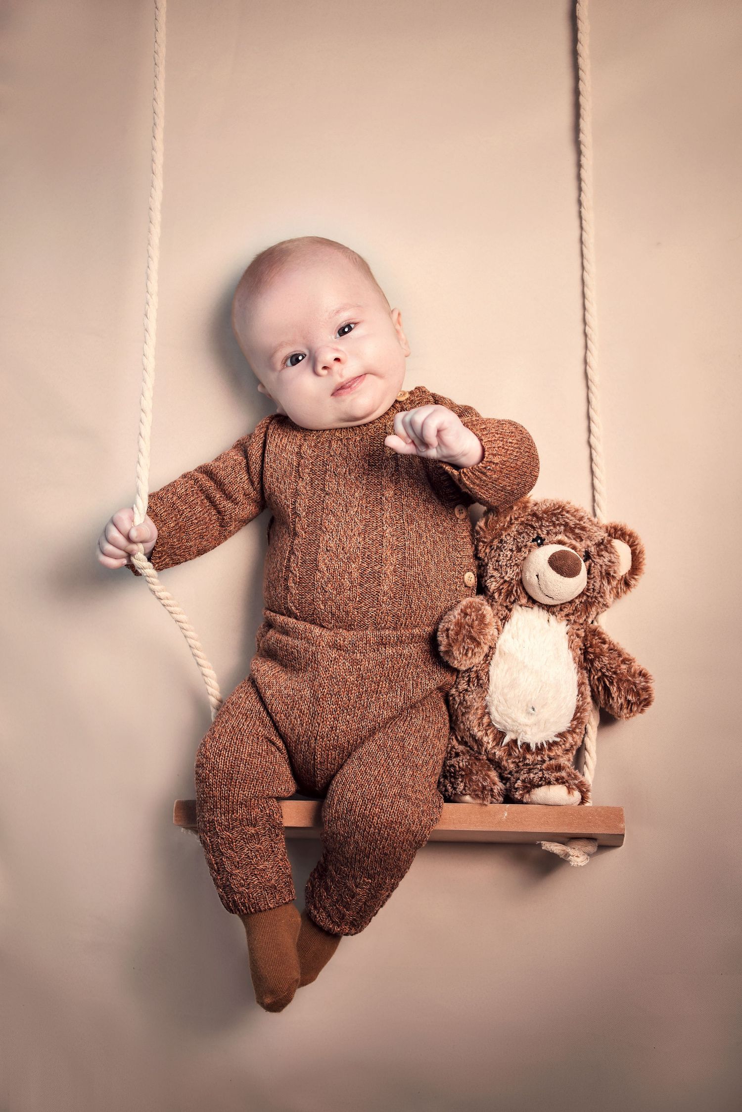 A baby in a brown onesie cuddles with a matching teddy bear on a soft neutral backdrop.