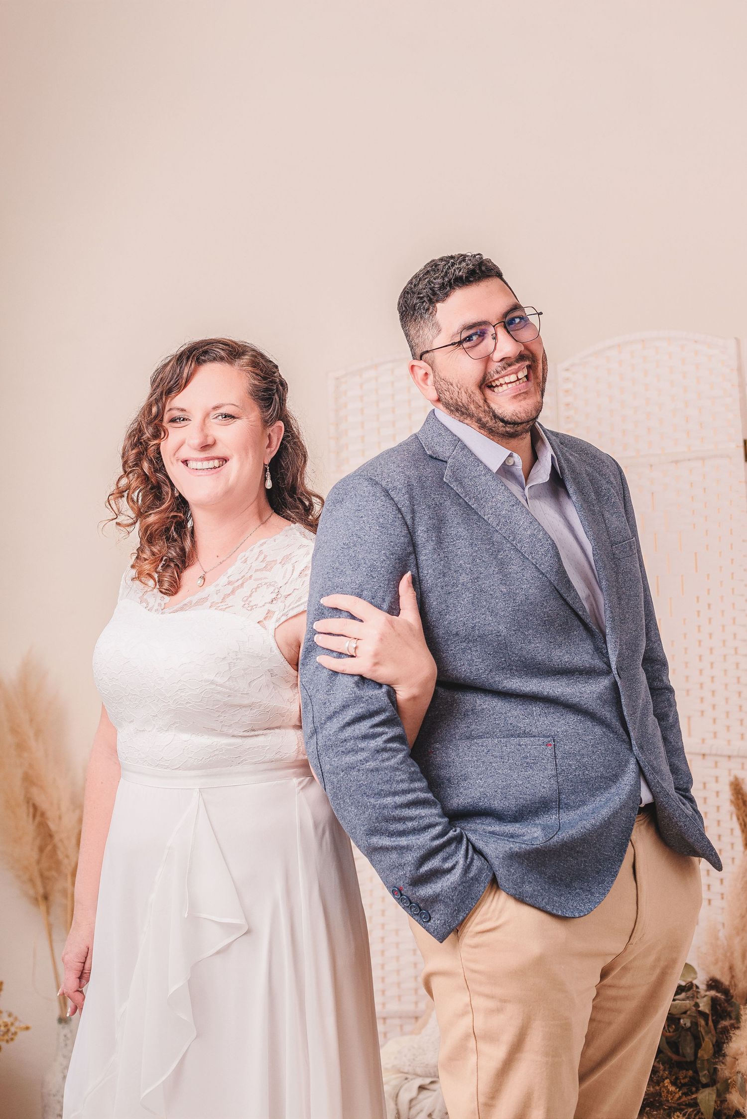 Playful couple in white dress and gray blazer pose together against a light backdrop during engagement photo shoot.